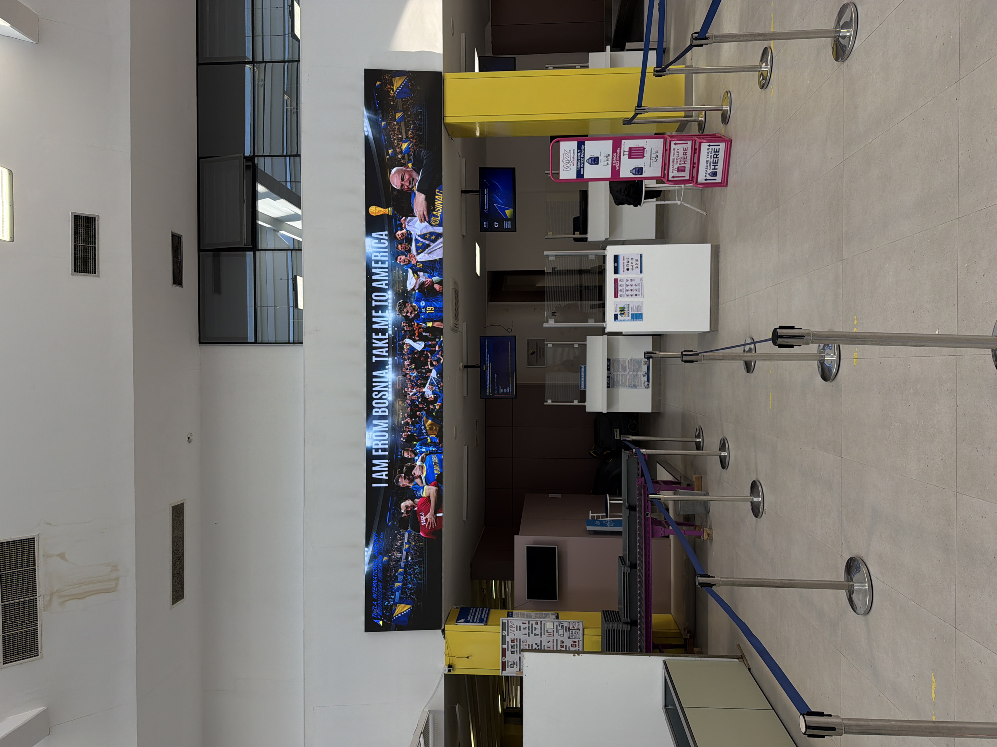 The image captures an interior view of the Tuzla International Airport in Gornje Dubrave, Bosnia and Herzegovina. A prominent feature is a large vertical banner that displays "I AM FROM BOSNIA. TAKE ME TO AMERICA" along with images of Bosnian athletes and supporters. Adjacent to the banner are various airport service counters in yellow and white, displaying information such as baggage size requirements. The queuing system is delineated by silver stanchions with blue retractable belts. The background reveals a modern airport interior with windows and structural elements. The floor is tiled and appears clean. The lighting is bright, possibly daylight enhanced by indoor lighting.