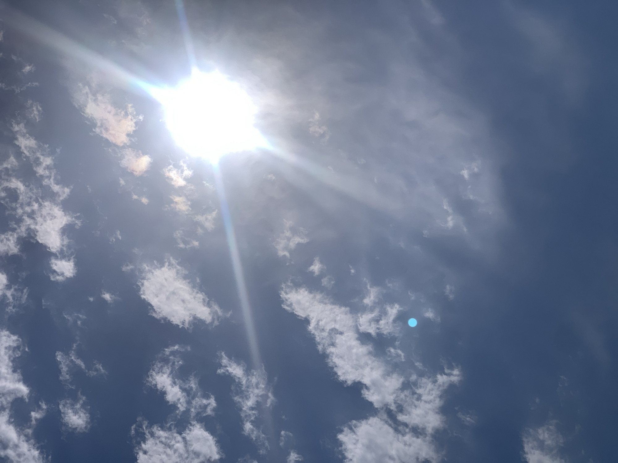 The image shows a bright sun with rays shining through scattered white clouds against a deep blue sky. There is a small, bright blue circular artifact in the lower left quadrant of the image. The sun is prominently positioned in the upper right of the frame. The scene appears to be an outdoor view of the sky, likely taken from ground level or a slightly elevated position, with no identifiable structures or elements of the city of Khazar Raion, Azerbaijan visible.