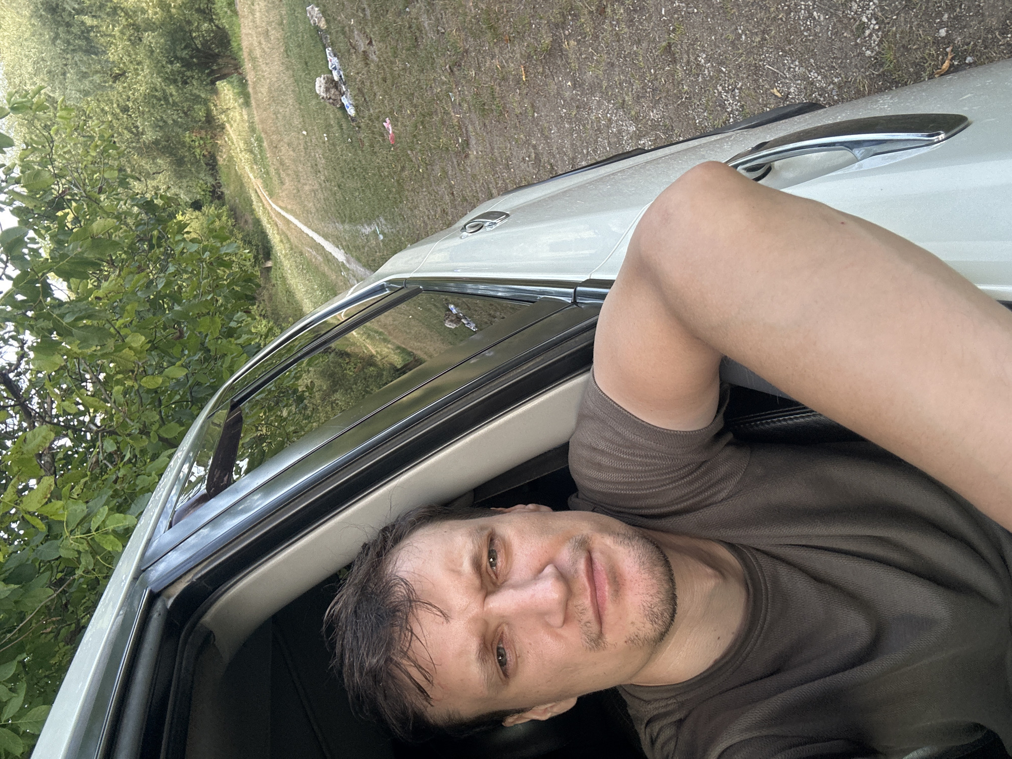 A close-up, eye-level, outdoor, natural light, selfie shows a man with short, dark brown hair and a light beard, wearing a dark olive t-shirt, leaning out of the driver's side window of a white SUV. The man is looking directly at the camera with a neutral expression, his left arm resting on the car door frame. Behind him, to the right of the vehicle, is a gravel road or path leading uphill, bordered by dry grass and some scattered debris. Lush green trees and foliage form the background. The image appears to have been taken in a rural or semi-rural setting, possibly on the outskirts of Makiv, Ukraine, given the presence of natural landscape elements consistent with the region.
