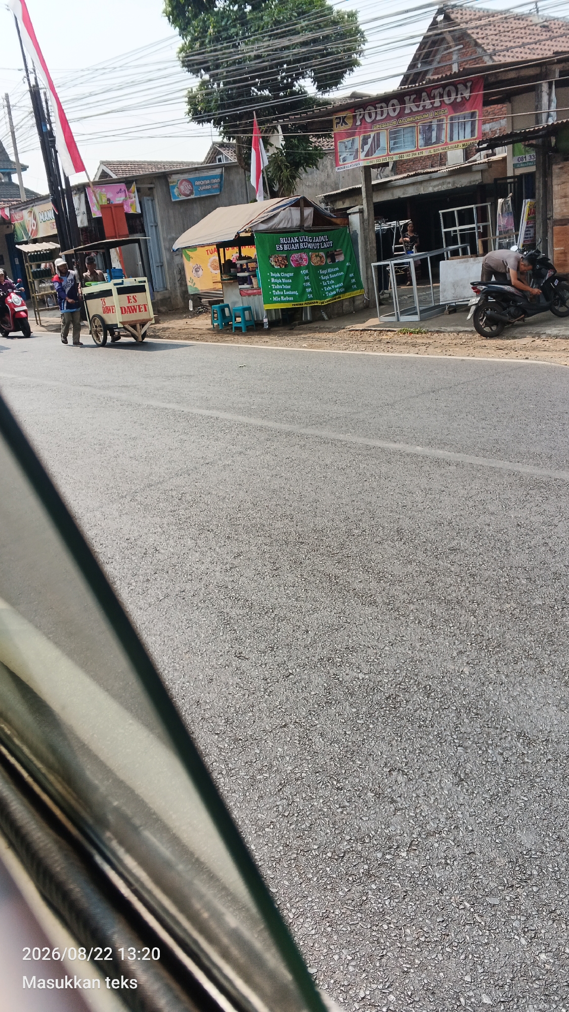 The image captures a street scene in Pakis, Indonesia, likely on August 22, 2026, at 13:20. In the foreground, a portion of a vehicle's windshield and interior is visible on the left. The road takes up a significant portion of the lower half of the frame. On the right side of the road, a shop with a red banner reading "PK PODO KATON" is present, advertising aluminum and glass products. To the left of this shop, a smaller stall with a green banner advertises "RUJAK ULIG JADUL ES BUAH RUMPUT LAUT," listing various menu items and prices. Several blue plastic stools are placed in front of this stall. Two individuals are visible near a mobile food cart labeled "ES DAWEET." One person appears to be pushing the cart while another stands nearby. A red motorcycle is parked on the far left side of the road, and a black motorcycle is parked in front of the shop on the right. The environment suggests a commercial area with shops and street vendors.
