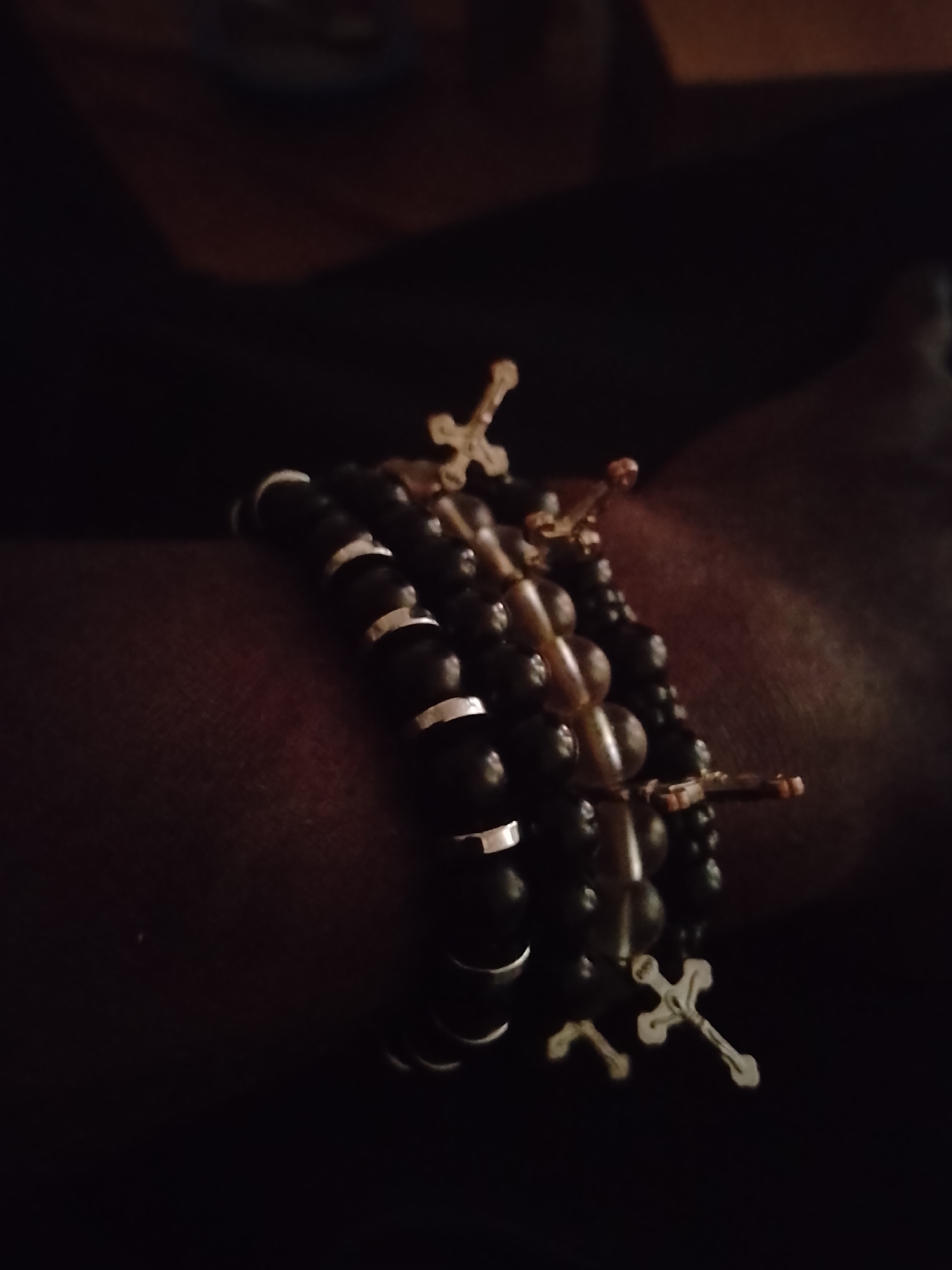 A close-up photograph depicts a dark-skinned arm adorned with multiple beaded bracelets. The bracelets are layered on the wrist, consisting of black beads with silver accents in one case and light brown beads in another. Several of the bracelets feature a silver cross charm. The image is taken in low light, creating a dimly lit environment. The location and city are unknown.