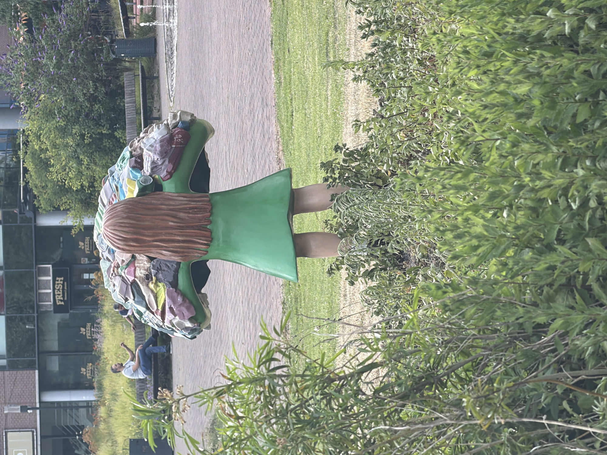 The image shows a life-sized statue of a young girl with her back to the camera, wearing a green dress. The top of her head is adorned with a cluster of colorful, bag-like objects, resembling groceries or shopping bags. The statue is situated in an outdoor urban setting, in front of a building with storefronts, one of which has a sign that reads "FRESH". A man is seated on a bench in the background, facing away from the statue and towards the building. Another person is partially visible on the same bench. The area around the statue is landscaped with green grass and various plants. In the further background, a fountain is visible, along with benches and trees. The overall scene depicts a public space with public art, suggesting a commercial or recreational area in a city. This location is the Julianapark in Rijswijk, Netherlands, where the statue "The Girl with the Shopping Bags" by artist Saskia van Vianen is located.