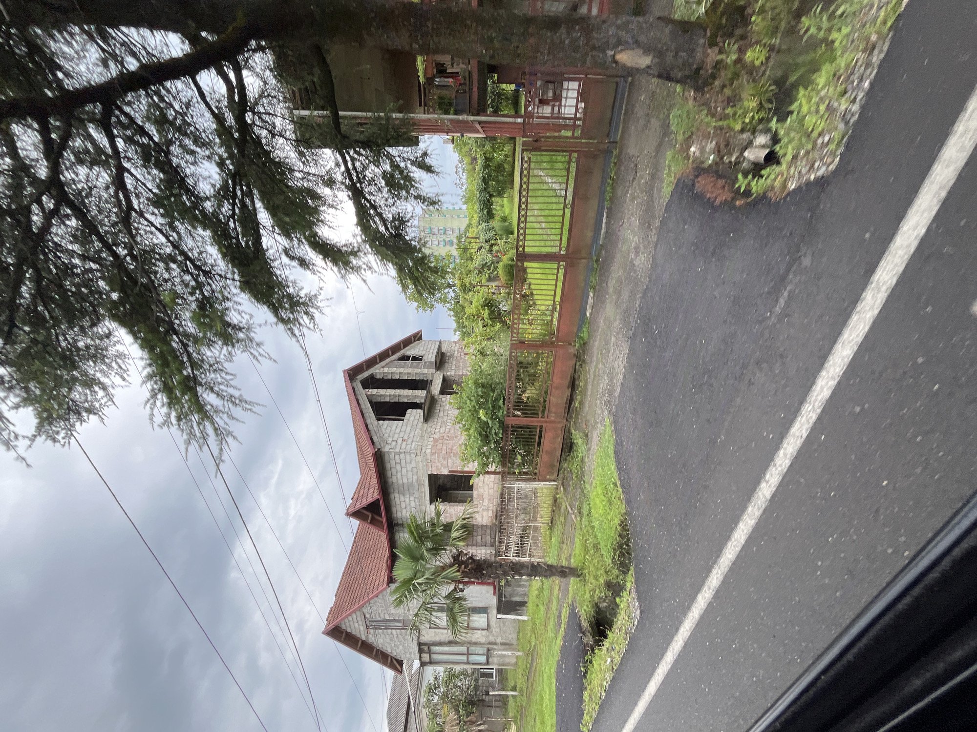 The image depicts a street view in Poti, Georgia, featuring a partially constructed building with a red tiled roof and cinder block walls. A small palm tree stands in front of the building. To the right, a rusted metal gate opens to a grassy yard with a large tree and other vegetation. In the background, a multi-story building is visible. The foreground shows a paved road with a white line marking. The sky is overcast and cloudy, suggesting it might be a damp or rainy day. The scene appears to be residential, with evidence of ongoing construction and overgrown greenery.