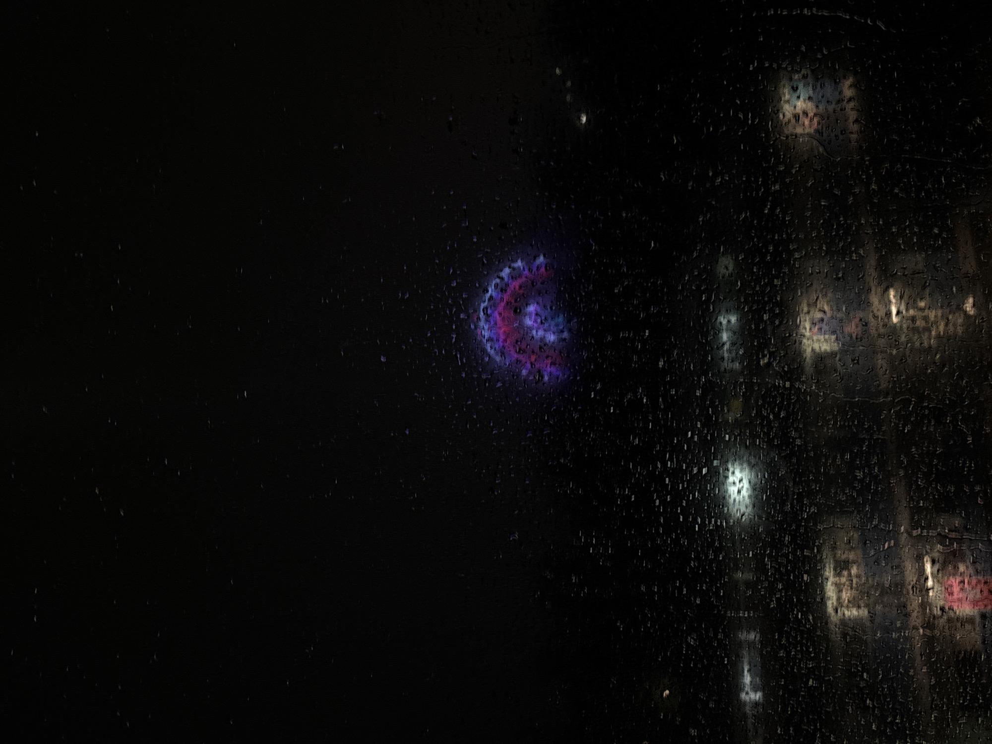 The image shows a rain-streaked window at night. Through the droplets, the blurred lights of a building can be seen. A circular, purple and pink light source is visible in the distance. Based on the available information, the precise location within Mexico City cannot be determined.