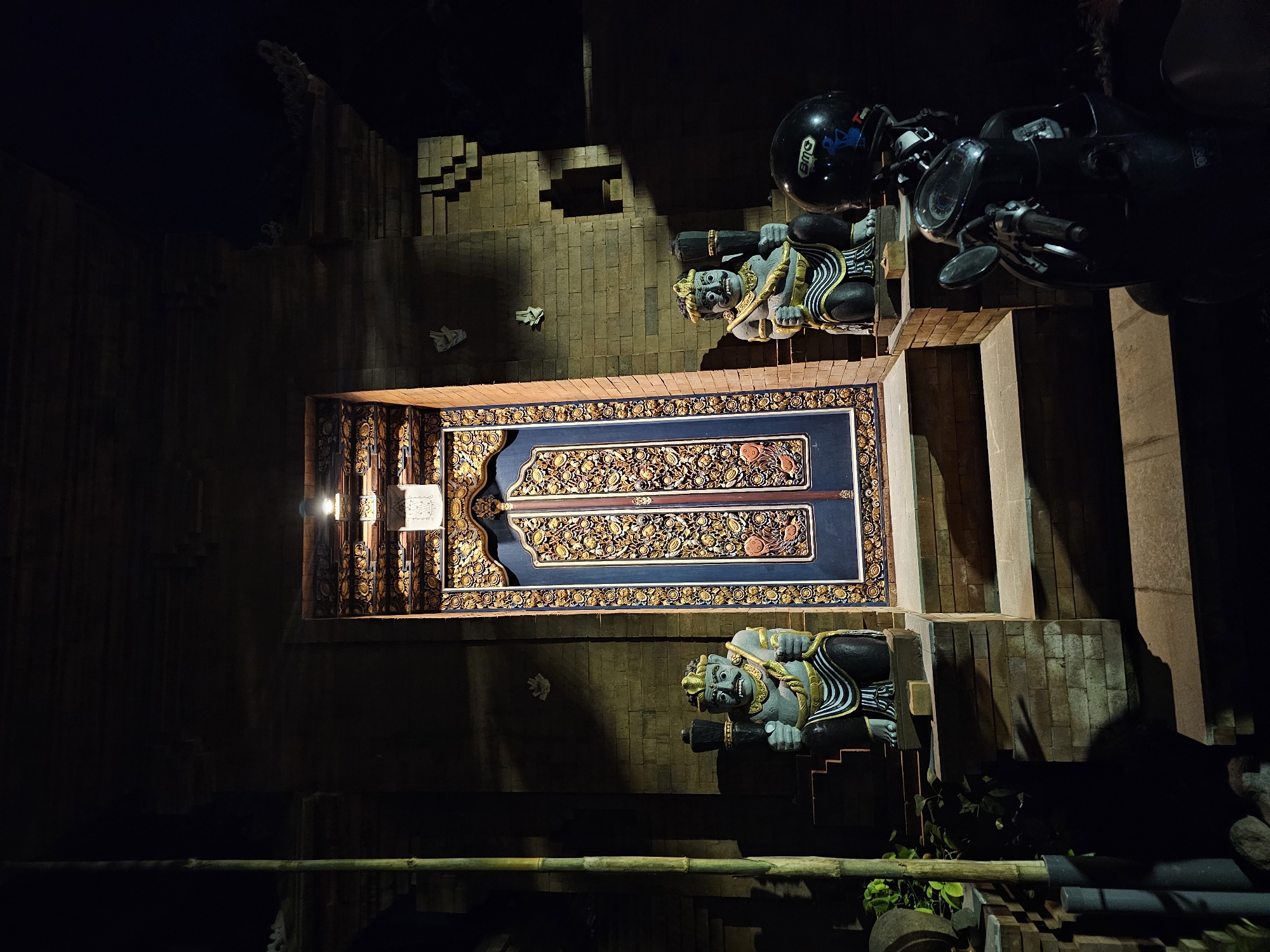 The image shows a nighttime scene outside what appears to be a temple or traditional building in Denpasar, Indonesia. In the center is a large, intricately carved double door, possibly made of wood, with blue and gold detailing. Flanking the entrance are two stone statues of guardian figures, each holding a club-like object. These figures are dressed in traditional attire with distinctive headdresses and facial features. In the foreground, to the right, a dark-colored scooter is parked, with a black helmet resting on its handlebars. The building is constructed of brick, and some bamboo poles are visible to the left of the frame, suggesting a tropical or subtropical environment. The lighting is dim, with a single light fixture illuminating the area above the doorway, casting shadows.