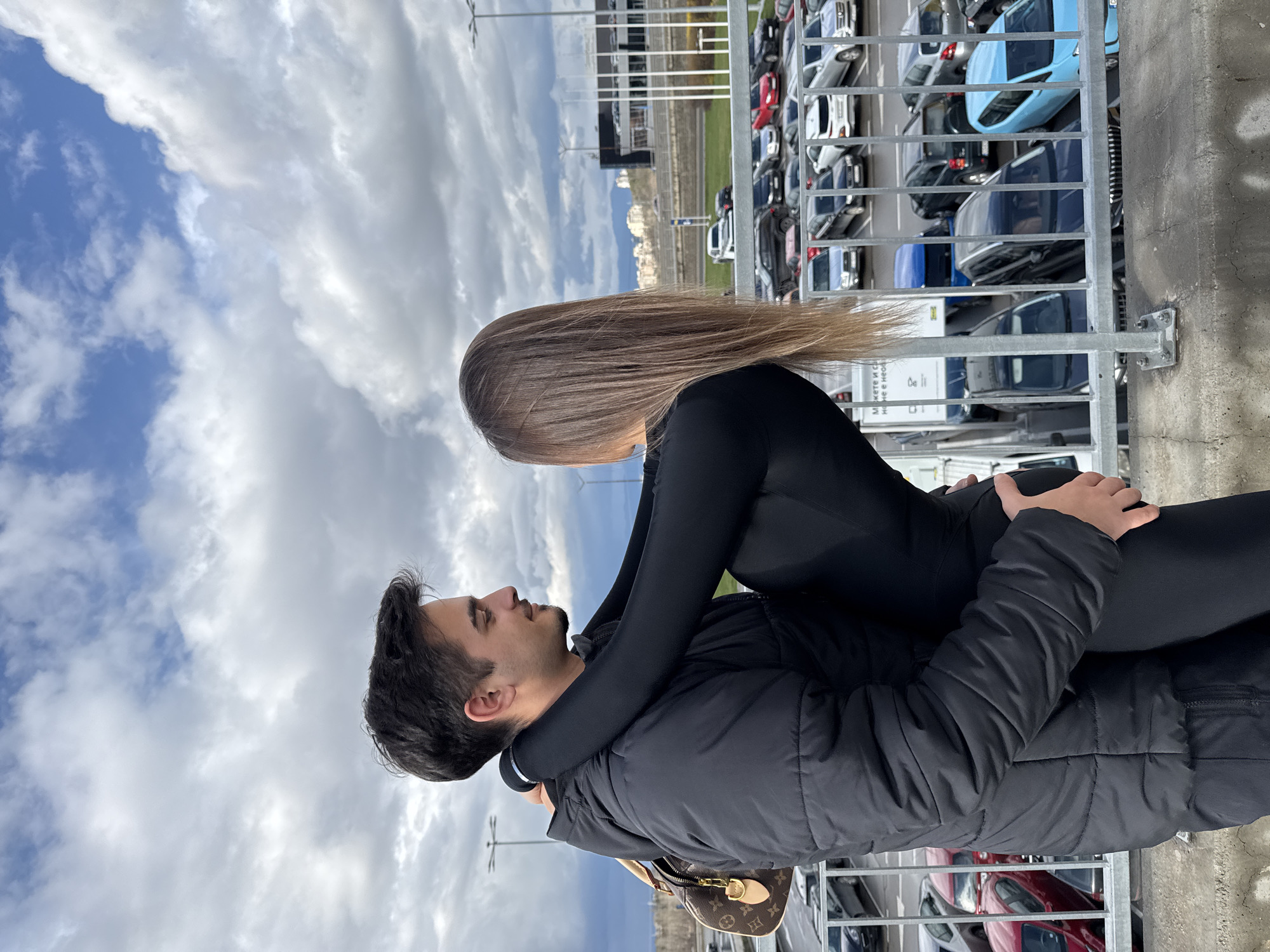 A man is carrying a woman in his arms in an outdoor setting. The man has short, dark hair, a dark jacket, and a Louis Vuitton handbag hanging off his shoulder. The woman has long, light brown hair and is wearing a black bodysuit. They are embracing. In the background, there is a parking lot filled with various cars, a building, and a sky with clouds. The photo was taken outside of the "Ring Mall" in Sofia, Bulgaria. The photograph is taken from a low angle.