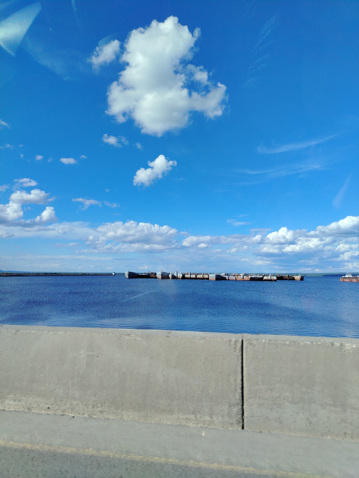 The image shows a view from a vehicle, likely a car, looking out over a body of water towards what appears to be a lumber storage area or shipping facility in Chaikovsky, Russia. A concrete barrier is visible in the foreground. In the middle ground, a large body of water reflects the bright blue sky dotted with cumulus clouds. On the water, long, narrow, dark-colored structures, likely rafts of lumber or similar materials, are visible. A single, larger ship is visible on the right side of the image, off in the distance. The sky is a deep blue with scattered white clouds.