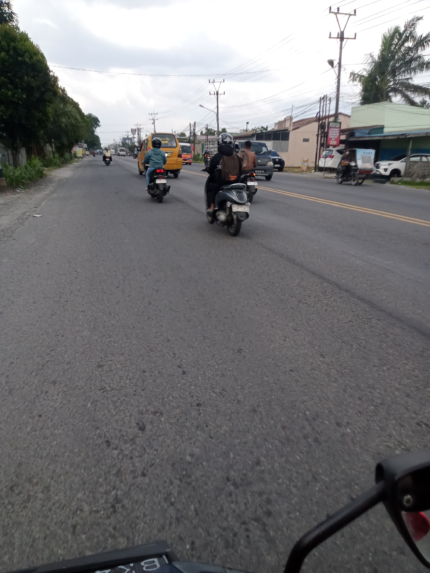 The image captures a view from the perspective of a motorcycle rider traveling on a two-lane asphalt road in Sei Semayang, Indonesia. The road has yellow double lines in the center, indicating a divided lane of traffic. On the left side of the road, there is a strip of green foliage and trees. On the right side of the road, there are buildings and power lines. The sky is overcast with clouds.
Several vehicles are visible on the road. In the foreground, a black motorcycle is partially visible at the bottom of the frame, and another black scooter with a rider wearing a helmet and backpack is moving away from the camera. Further down the road, a yellow van is traveling in the same direction. Beyond the van, a dark-colored car is also in motion. On the right side of the road, a motorcycle with a sidecar is carrying cargo and moving away from the camera, and two white cars are parked or moving slowly.
The scene depicts a typical day with moderate traffic on a highway or main road. The lighting suggests it is daytime, but the overcast sky creates a diffused light.