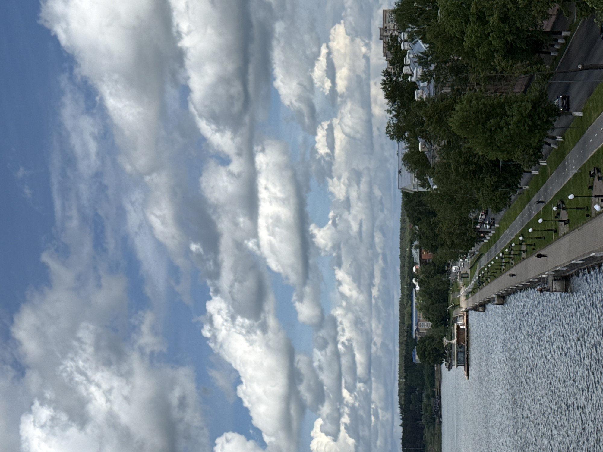 The image depicts a section of Votkinsk, Russia, with a significant focus on the sky filled with cumulus clouds. The lower portion features a body of water, likely a lake or reservoir, with a promenade lining the edge. The promenade has decorative lamp posts along its length. A few figures can be seen walking on the promenade. On the other side of the promenade, there are trees and buildings, including a multi-story structure. A road runs parallel to the promenade, and there are vehicles visible on the road.