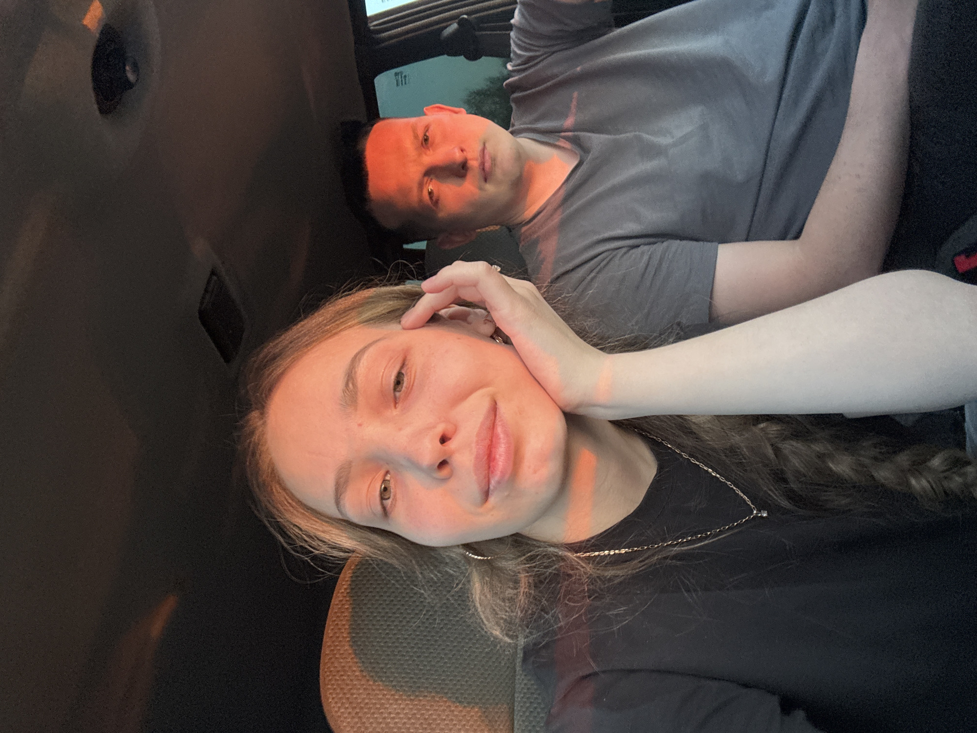 A selfie shows a young woman with long, wavy hair, wearing a black shirt and a silver necklace, leaning her head on her hand. Behind her is a man wearing a grey t-shirt. They appear to be inside a vehicle. The image is tilted. There is no information available to confirm the location is in Ivanovka, Russia.