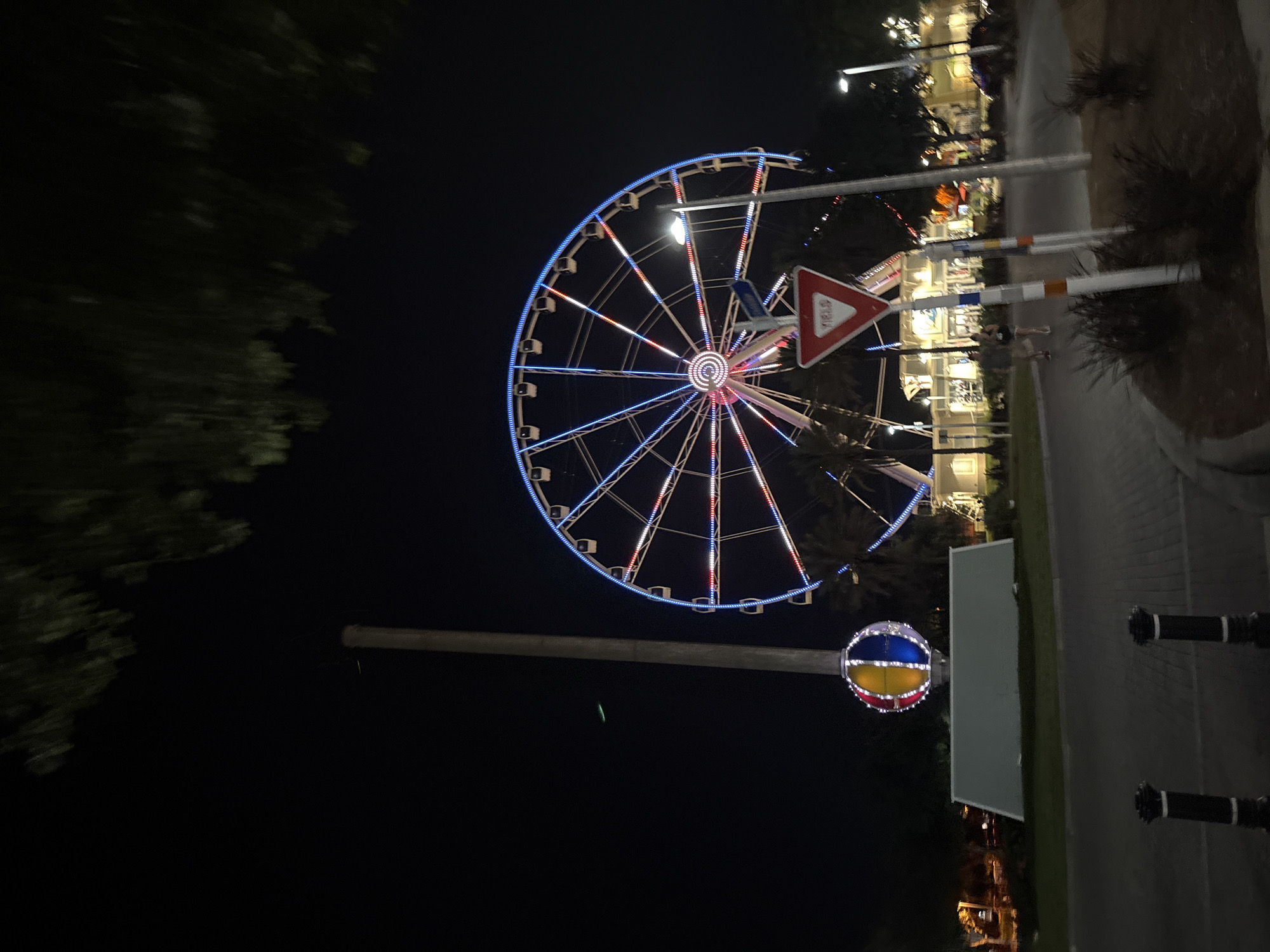 The image depicts a nighttime scene in Panama City Beach, Florida, USA, showcasing a large Ferris wheel illuminated with blue and red lights. The Ferris wheel is the central focus, with its intricate structure and vibrant lights dominating the frame. To the left of the Ferris wheel, a tall pole stands with a spherical, multicolored decoration at its base, also lit up. In the foreground, a paved walkway leads towards the Ferris wheel. A "YIELD" sign is visible, positioned near a street light and some vegetation. In the background, there are buildings that appear to be part of an entertainment venue or shops, with some lights on, suggesting activity. Several people are visible walking on the paved area in front of the buildings, seemingly enjoying the evening. The overall environment suggests a lively tourist or amusement area. A tree with dark foliage is visible at the top of the frame, casting shadows and adding to the nighttime ambiance. The sky is dark, typical of nighttime.