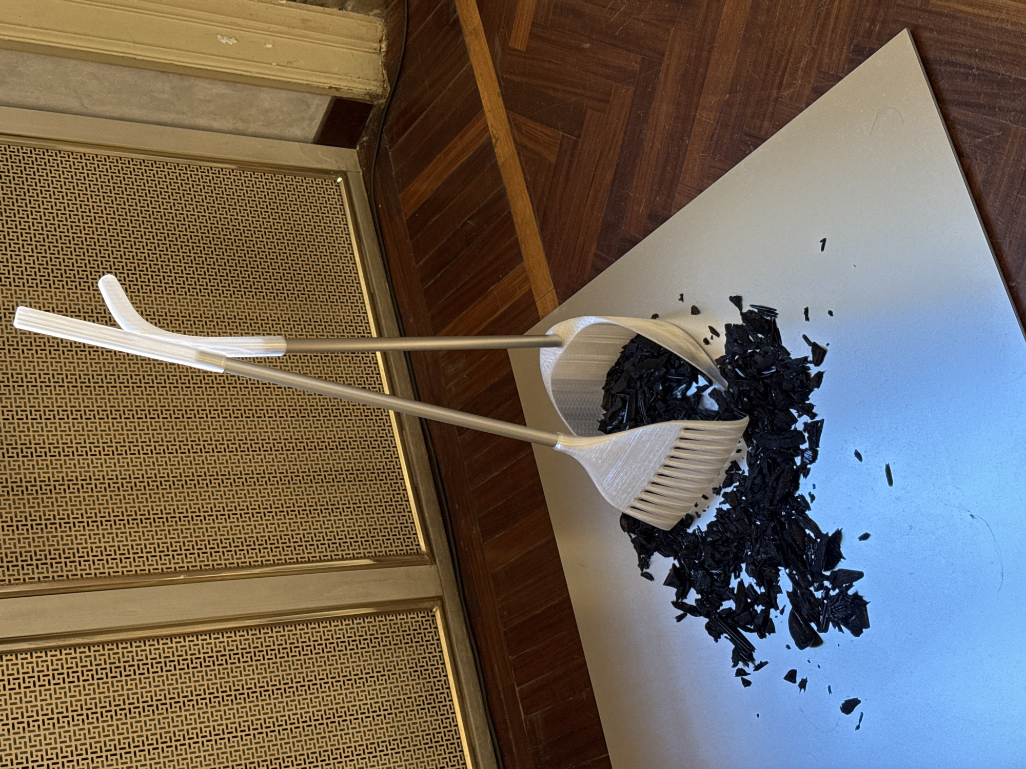 A dustpan and brush set sits atop a pile of what appears to be broken black plastic, spread across a light gray surface. Adjacent to this, a patterned metal grate with a gold frame is partially visible. To the right of the grate, a parquet wood floor is seen. The scene appears to be indoors, but no definitive markers point directly to Milan, Italy. There are no people present in the frame.