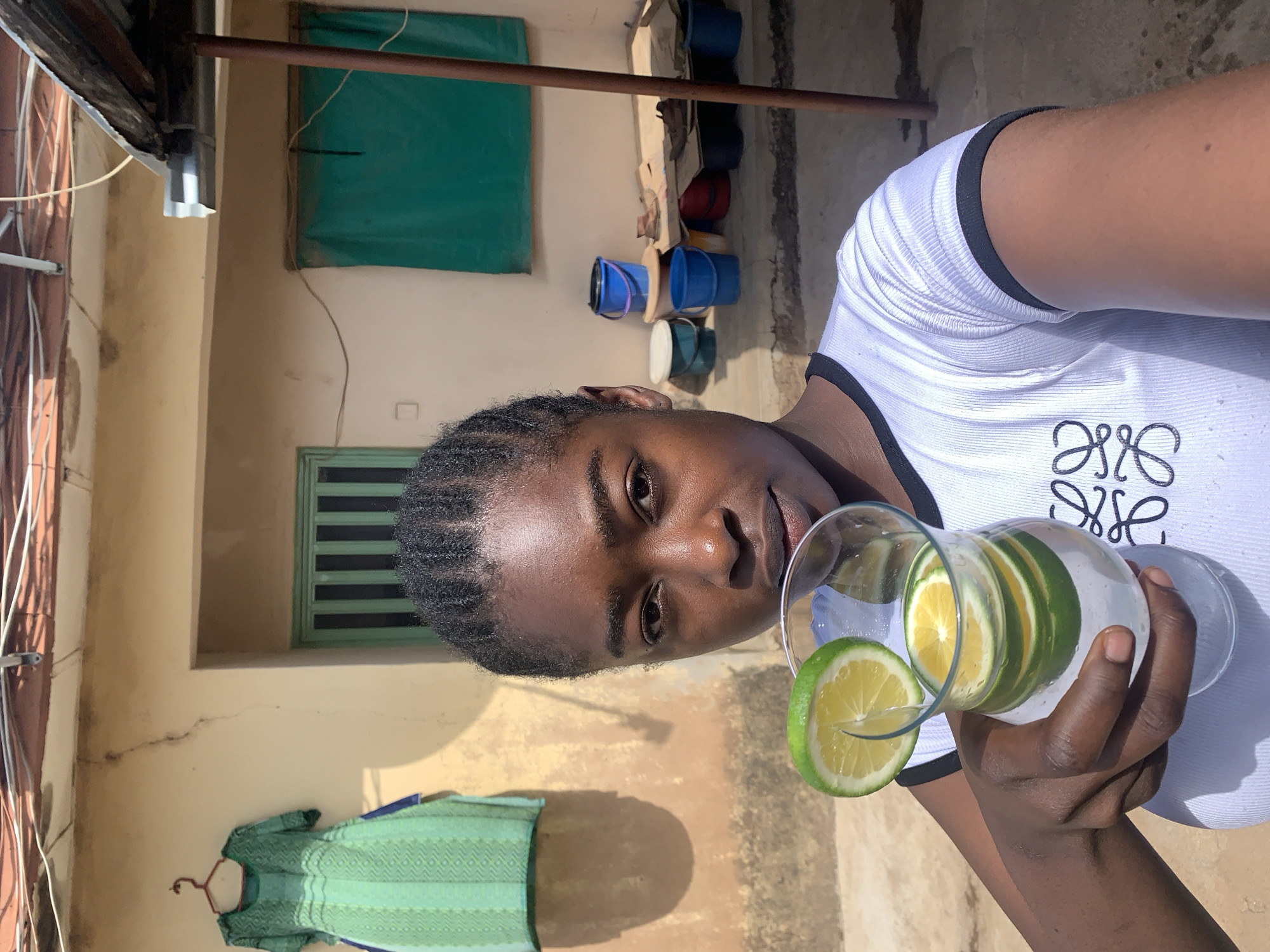 A person with dark skin and braided hair holds a glass pitcher filled with water and sliced lemons. The person wears a white shirt with black trim and a graphic design on the front. The setting appears to be outdoors near a building with a pale yellow wall. A green dress hangs on a hanger against the wall. Other details include a barred window, various buckets stacked on the ground, and a piece of green fabric hanging over another window. The city is Rafin Par, Nigeria.
