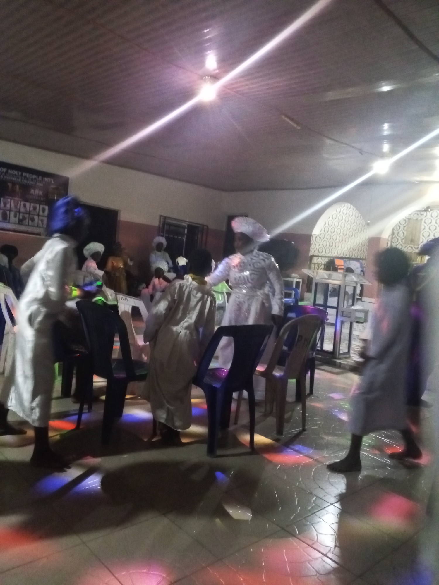 In a dimly lit interior, people are gathered, some standing and some sitting on chairs. The individuals are dressed in white robes and head coverings. The floor is tiled and illuminated with colorful spotlights, casting patterns on the surface. Overhead lighting is visible on the ceiling. A poster hangs on the wall, and a podium-like structure is present in the background. The location is in the city of Unknown, Unknown.