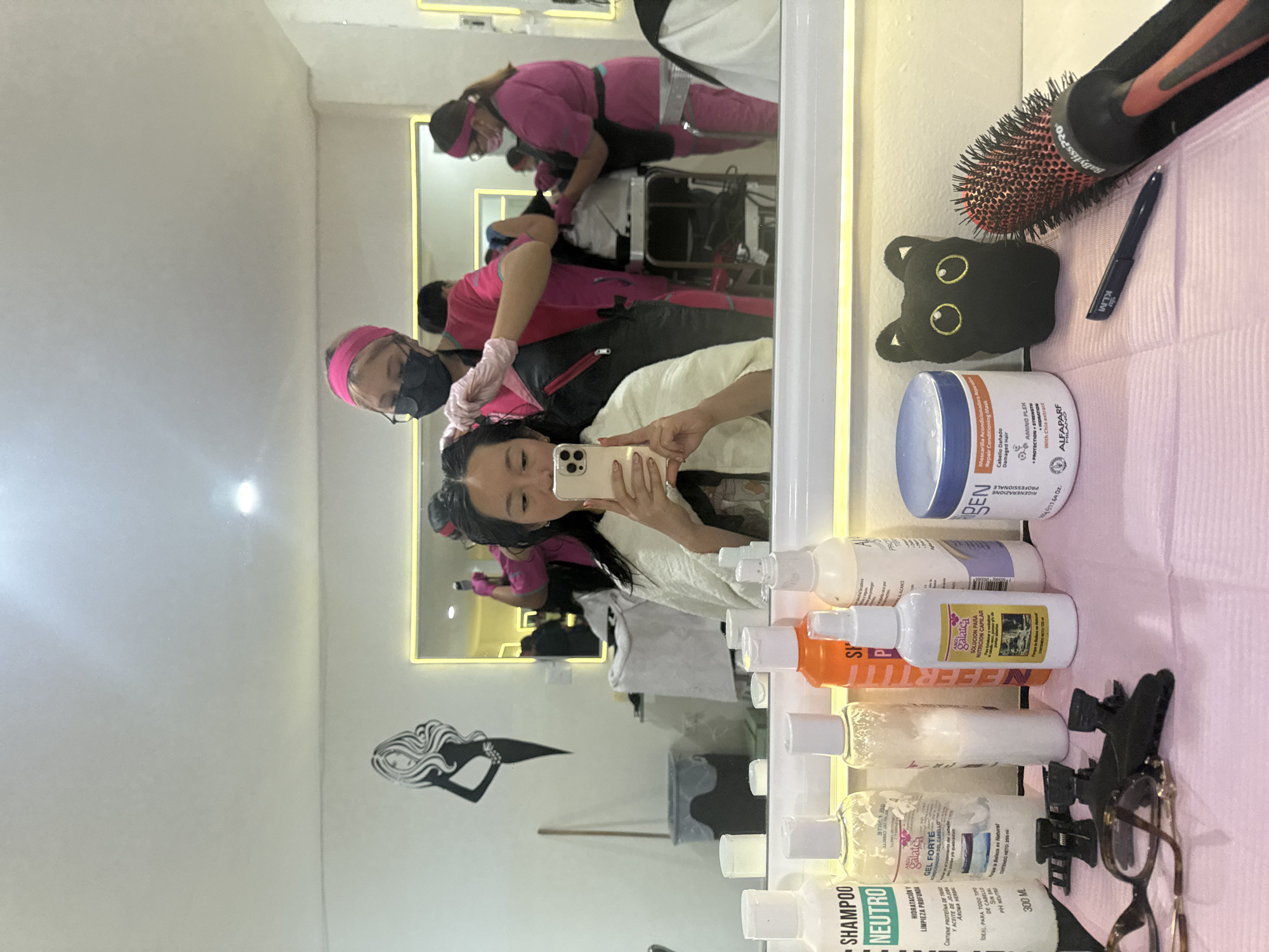 The image, rotated 90 degrees counter-clockwise, captures a mirror selfie taken inside a hair salon in Puebla, Mexico. The foreground is dominated by various hair products and tools neatly arranged on a pink surface next to a mirror. These include a large orange and black BaBylissPRO hairbrush, a black cat-shaped eye mask with yellow eyes, and a small dark blue razor or pen with "KLIM" visible. Several bottles of hair products are lined up, along with hair clips and a pair of eyeglasses.
Reflected in the mirror, the person taking the selfie is visible from the chest up, holding a light-colored smartphone. Their hair appears damp, suggesting a treatment in progress. Behind them, also reflected, are two salon staff members wearing pink uniforms or scrubs with matching headbands. One staff member, wearing a black face mask and glasses, is actively working on the customer's hair. Further back, another staff member in pink is also visible. The salon walls are light-colored, and a black and white mermaid decal adorns one wall. Bright lighting strips are visible in the reflection.
Visible text on the products includes:
* A "SHAMPOO NEUTRO" bottle mentioning "HIDRATACIÓN Y LIMPIEZA PROFUNDA," "PROTEÍNA DE TRIGO Y ACEITE DE JOJOBA," "AROMA HERBAL," "IDEAL PARA TODO TIPO DE CABELLO," "SIN SAL," "PH NEUTRO," and "300 ML."
* A "galatca GEL FORTE" bottle.
* A partially visible bottle with text like "CIONADOR DEL CABELLO quebradizo CONTENIDO NETO: 250 ml."
* A bottle labeled "NEFERTITI."
* A white bottle from "galate" for "SOLUCION PARA NUTRICION CAPILAR."
* A large blue and white jar with "BEN RIGENERAZIONE PROFESSIONALE 17.64 Oz." and "Mascarilla Acondicionadora Repair Conditioning Mask Cabello Dañado Damaged Hair AMINO PLEX PROTECTION STRENGTH HIDRATION With Chia extract ALFAPARF Milano."
* The hairbrush has "BaBylissPRO" printed on it.
* The razor/pen has "KLIM" printed on it.