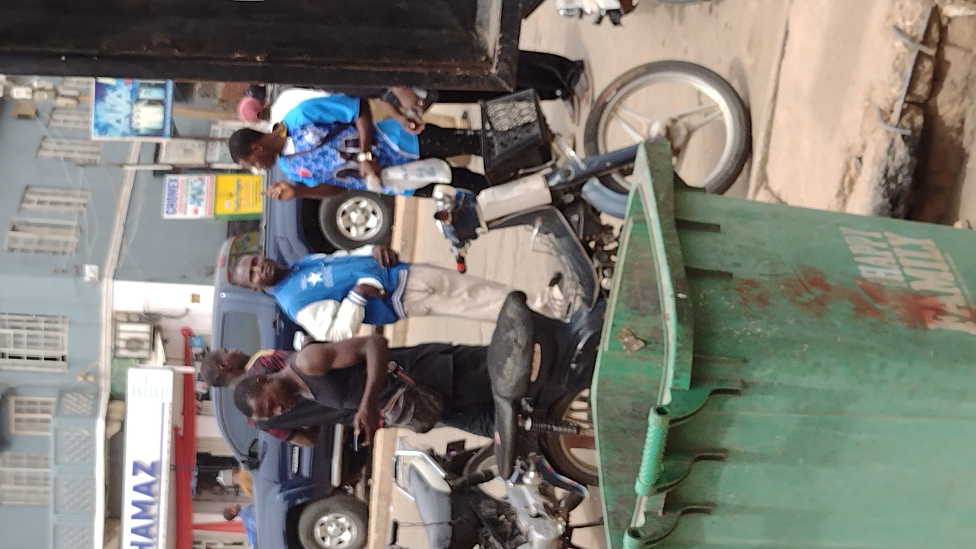 This image, originally rotated 90 degrees counter-clockwise, depicts a bustling street scene during daylight, likely in an urban area such as Jos, Nigeria. In the foreground, a large, dark green utility container or refuse bin dominates the right side, with the white text "HAPPY MILY" clearly visible, suggesting it might be part of "HAPPY FAMILY."
Multiple men and vehicles populate the street. Towards the bottom center, two men are positioned near a dark-colored motorcycle with a black seat. One man wears a black tank top, and the other a black t-shirt, appearing to be engaged in a casual interaction or inspecting something. Further into the frame, a man in a blue and white varsity-style jacket stands beside a dark blue SUV or truck, looking towards the viewer's right. Above him, another man, dressed in a blue patterned shirt with white details, is seen interacting with someone out of frame or gesturing with his hand.
In the background, multi-story buildings with numerous windows line the street. Several commercial signs are visible, including a prominent yellow sign displaying "CHROMEX" and a white signboard that reads "HAMAZ." The front wheel of another motorcycle is also partially visible near the top right. The overall atmosphere is that of a typical, active street during bright, clear daytime conditions, with no apparent signs of adverse weather. The activity suggests a routine gathering or daily life on the street.