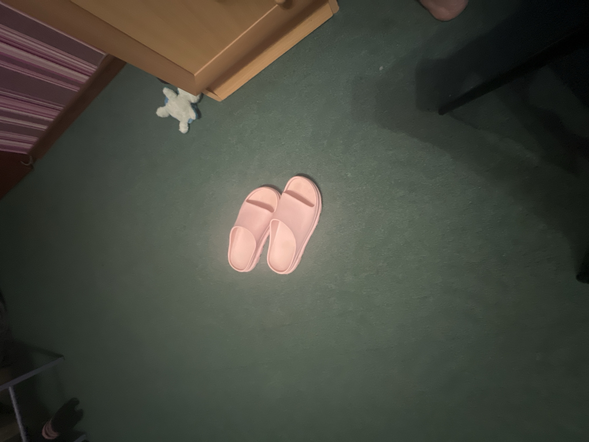 This is a close-up, high-angle shot of a pair of pink slip-on sandals on a dark green carpet. The sandals are positioned in the center of the frame, with one slightly in front of the other. To the right, a portion of a light-colored wooden dresser is visible, with some pink and white striped fabric draped over its top edge. A small, light blue plush toy is also visible near the dresser. The scene appears to be indoors, likely a bedroom or living area. There is no indication of the specific location within the city of Saratov, Russia.