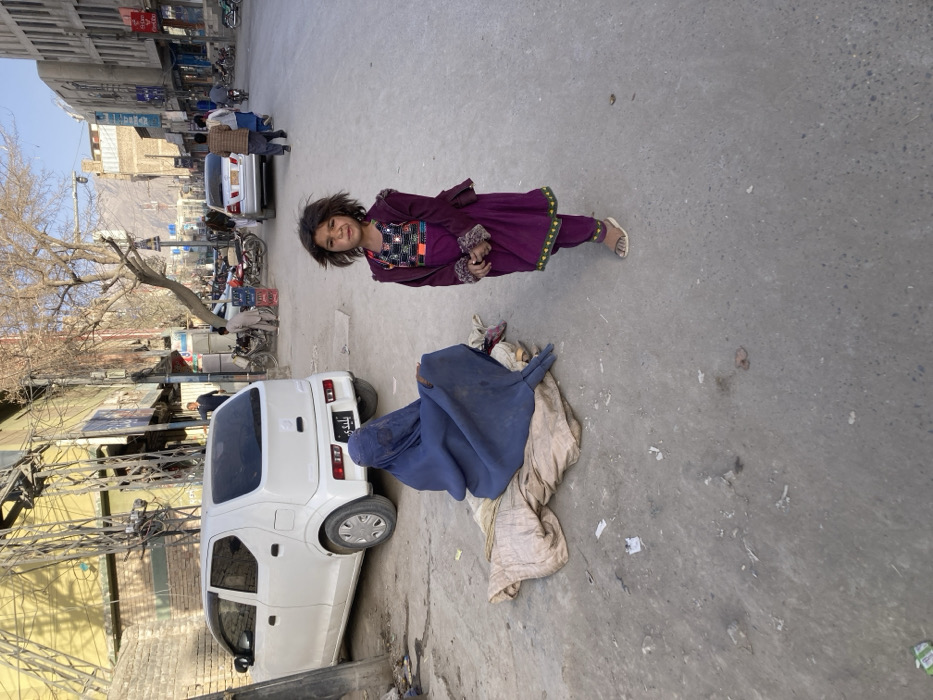 This image depicts a street scene in Quetta City Tehsil, Pakistan, under natural daylight. In the foreground, a young child, approximately 5-7 years old, stands slightly right of center, facing the camera. The child has dark hair, a dark complexion, and is dressed in a long-sleeved purple top with a black and colorful embroidered chest panel, purple pants with green trim, and light-colored sandals. To the child's left, an adult figure sits directly on the worn concrete pavement, covered completely in a dark blue burqa and positioned on a light beige fabric. The ground around the subjects is dirty and littered with small pieces of debris.
In the midground, a white Suzuki Alto hatchback is parked on the left, facing away from the camera; its rear license plate contains the Urdu/Farsi script "بلدی" (Baladi/Municipality). Beyond it, the street extends into the background, populated by several male pedestrians and various vehicles, including multiple motorcycles parked on the right and a silver sedan further down the road. Buildings, approximately 2-3 stories high and exhibiting commercial signage, line both sides of the street. Overhead, complex utility wiring is visible on poles to the left. Bare, deciduous trees are intermittently placed along the thoroughfare. In the distant background, rugged mountains are visible under a clear, light blue sky.
