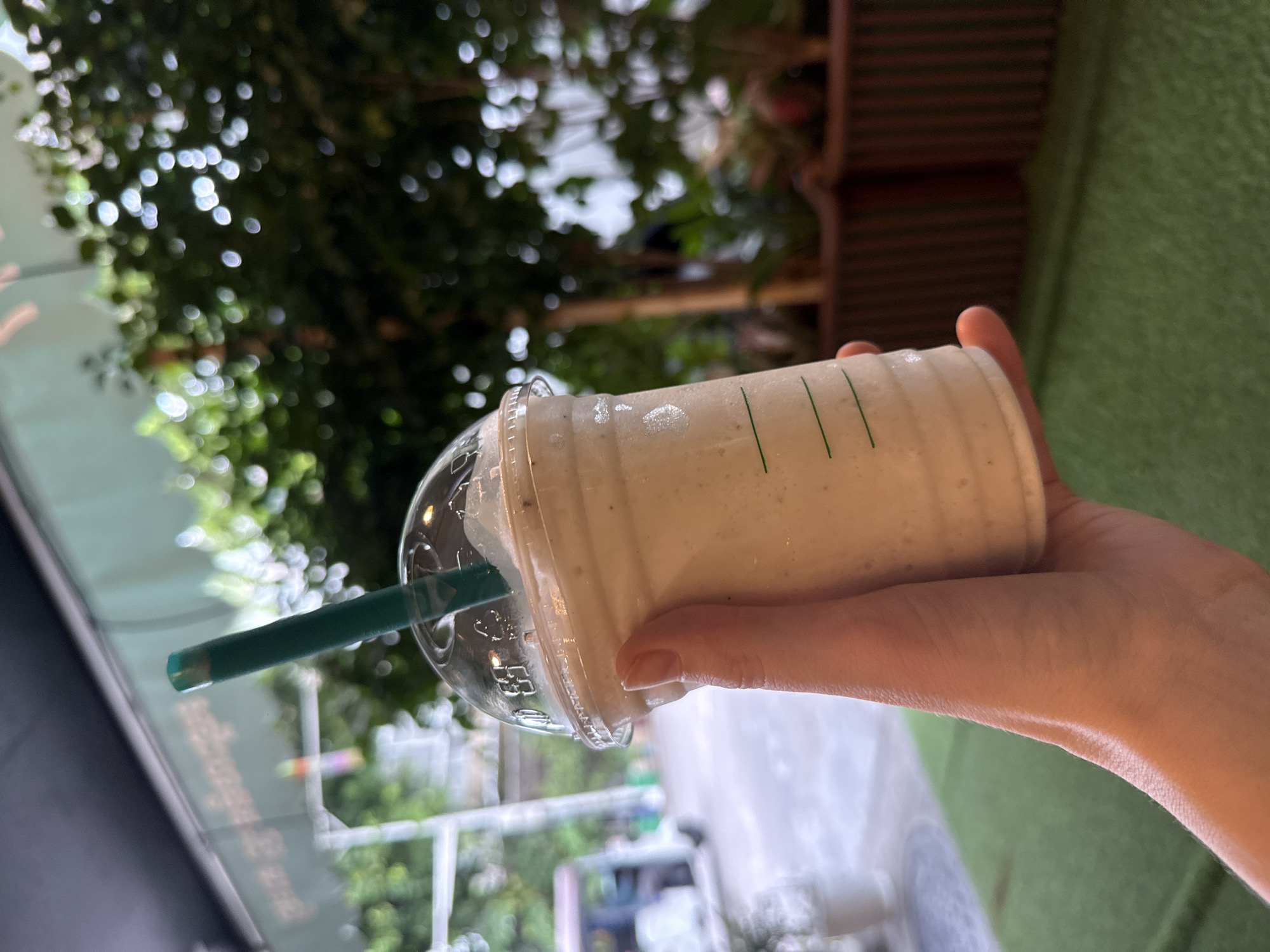 A person's hand holds a plastic cup filled with a light-colored, blended beverage. A green straw extends from the cup. The cup has green lines printed on it. In the background, out of focus, is a lush green foliage, some vertical brown striped planters, and a white vehicle. The scene appears to be outdoors, possibly in a cafe or restaurant setting in Khan Chbar Ampov, Cambodia, indicated by the style of the vehicle and the presence of greenery. The ground is covered with a green, textured material, possibly artificial turf.