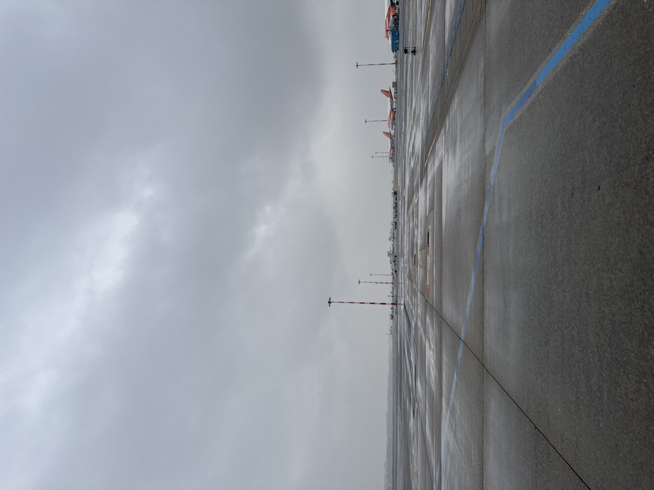 The image depicts an airport tarmac under an overcast, uniformly grey sky, indicating a dull, potentially wet day. The ground, a vast expanse of dark grey asphalt and concrete, is reflective with moisture, suggesting recent rain. A prominent blue curved line marks the foreground pavement, with additional white and faint red lines and rectangular panel divisions visible. In the mid-ground, a series of red and white striped poles extends diagonally into the distance. Several white ground support equipment vehicles are parked further back. The background features multiple commercial aircraft, predominantly orange-tailed and white-bodied, consistent with EasyJet livery. At least four such aircraft are identifiable, with a blue ground service vehicle positioned next to the nearest aircraft on the right. A low horizon line with indistinct distant shapes separates the tarmac from the sky. No individuals, specific actions, or interactions are visible. This scene is identified as Milan Malpensa Airport (MXP), situated in the city of Somma Lombardo, Italy.