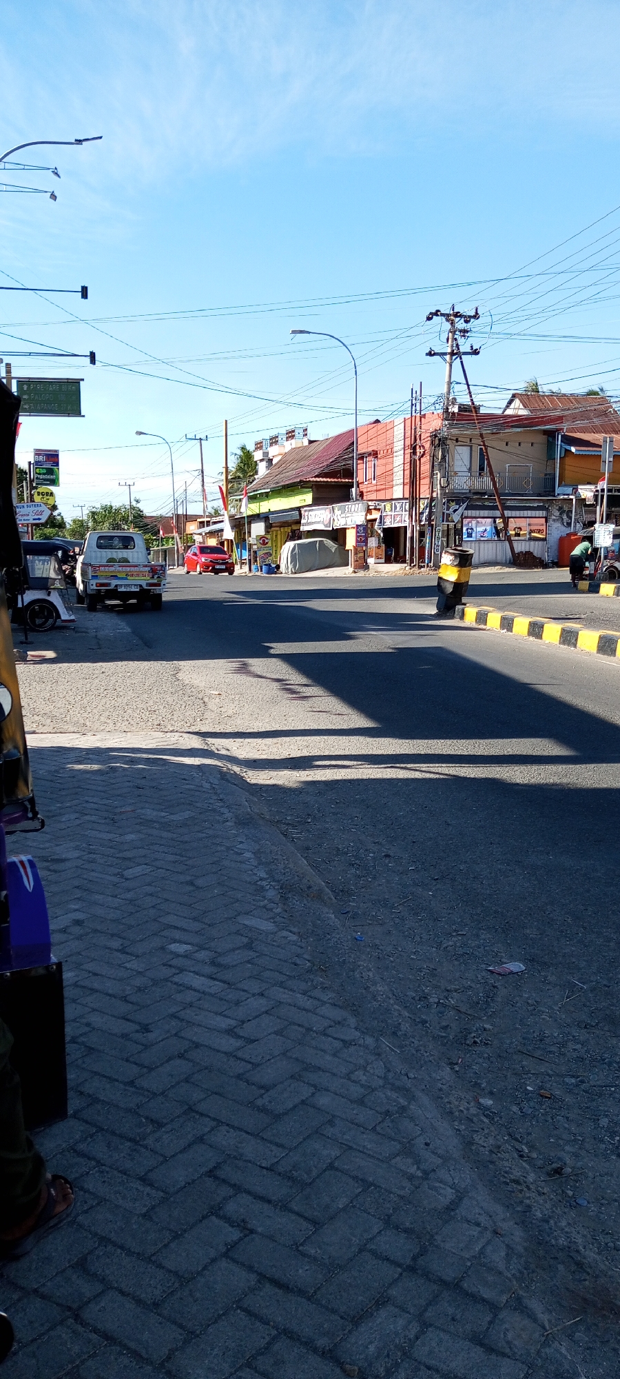 The image captures a street scene in Assorajang, Indonesia, on a clear day. The road is asphalt, bordered by a sidewalk made of grey interlocking bricks. On the right side of the road, there is a bright yellow and black striped barrier. Further down the street, buildings line the road, with shops and houses. Several vehicles are visible: a white pickup truck, a red car, and a three-wheeled vehicle on the left. Power lines and streetlights are present. A signpost with distances to different locations is visible in the upper left quadrant of the image. The overall environment appears to be a commercial or residential area within the city.