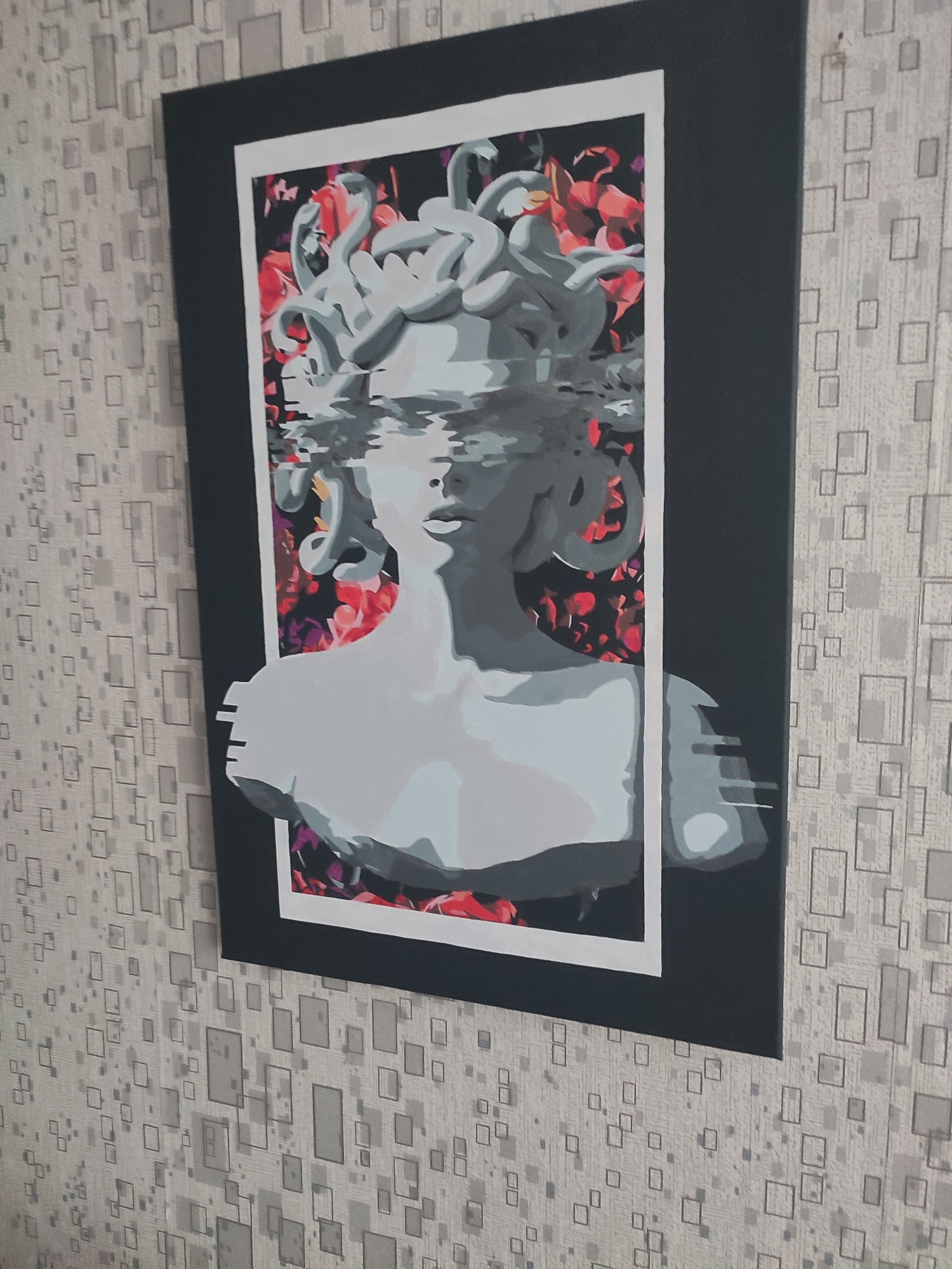 The image shows a framed painting hanging on a wall covered with patterned wallpaper. The painting depicts a stylized bust of Medusa with her snake-hair rendered in grey and white, and a glitch effect obscuring her eyes and part of her face. The background of the painting is a collage of red and black floral elements. The wallpaper has a light grey textured background with a repeating pattern of darker grey squares of various sizes. The painting is framed with a black matting and a thin white inner border. The image is taken from an upward angle, with the painting dominating the center. The location of the city of Narowlya, Belarus is not ascertainable from the visual information provided in the image.