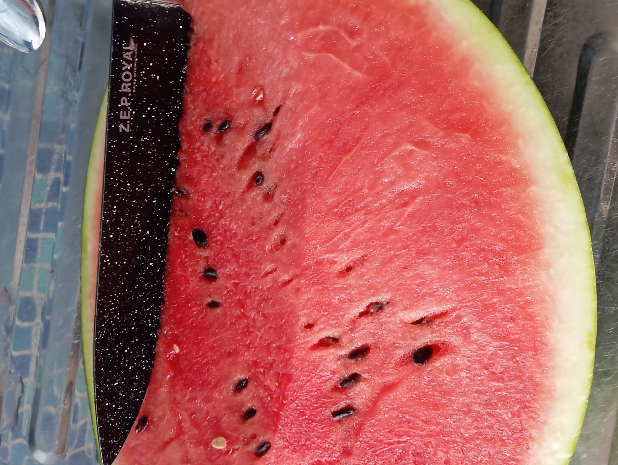A slice of watermelon is positioned on a grey, ridged surface, with a knife lying on top of the watermelon, partially obscuring it. The watermelon slice is vibrant pink with numerous black seeds scattered throughout its flesh and a pale green rind. The knife has a black speckled handle and blade, with the text "Z.E.P. ROYAL SWISS INTERNATIONAL" visible on the blade. The background consists of a textured, blue and green tiled surface. There is no information in the image to indicate a location in the city of Temryuk, Russia.