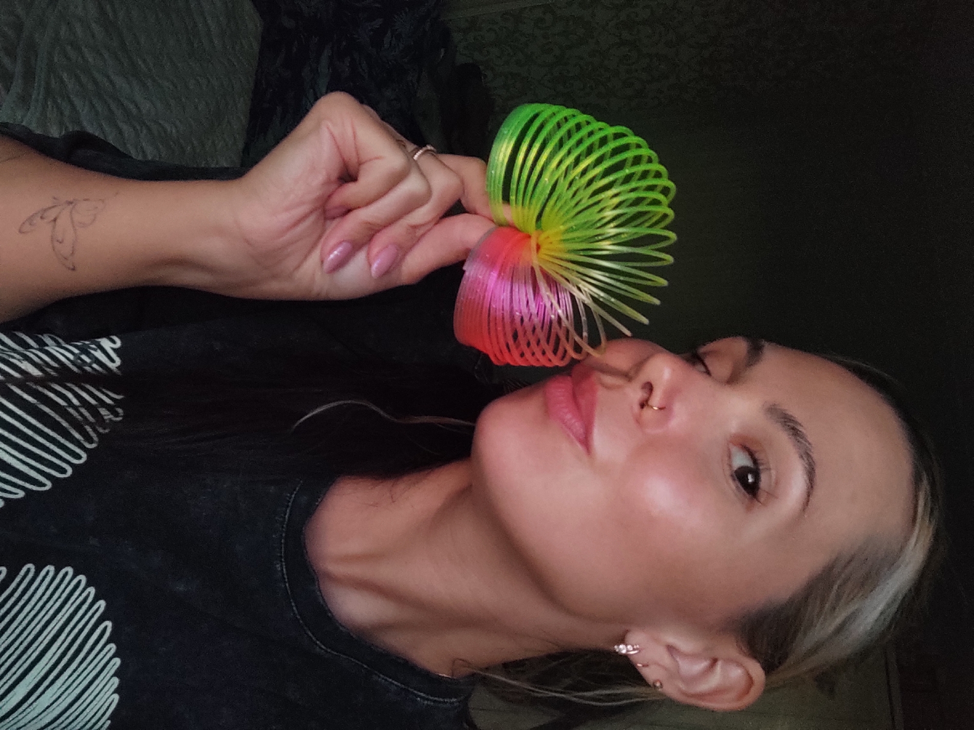 A woman is holding a rainbow-colored Slinky toy in front of her face. She has a nose ring and multiple piercings in her ear. The woman is wearing a black shirt with a white spiral design. She has a butterfly tattoo on her left wrist and a ring on her finger. The background is dark and indistinct. The location in the city of Ольховка, Russia, cannot be determined from the image.