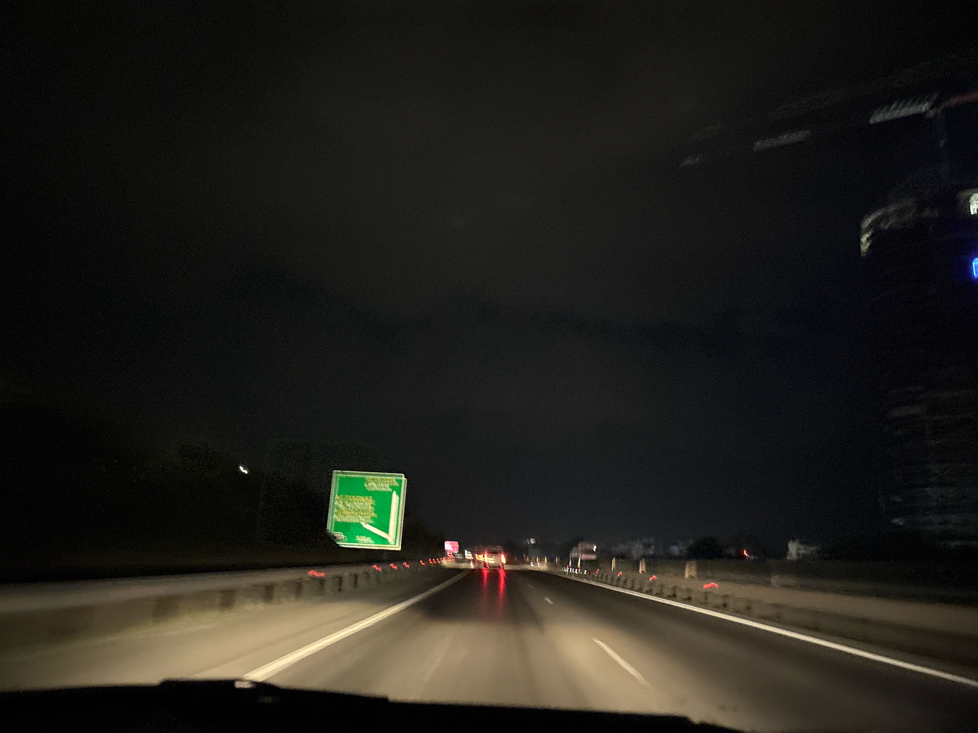 A first-person perspective shot from inside a vehicle at night on a highway. The road surface is wet and reflective, with lane markings visible. To the left, a green road sign with white text is illuminated. Along the side of the highway, orange traffic cones are placed at intervals. In the distance, other vehicles with illuminated headlights and taillights are visible, creating streaks of light due to motion blur. A tall, dark structure, possibly a building or a tower, is visible on the right side of the frame, partially illuminated. The sky is dark and overcast. The image appears to be taken on a highway in the Agios Tychona Community, Cyprus.