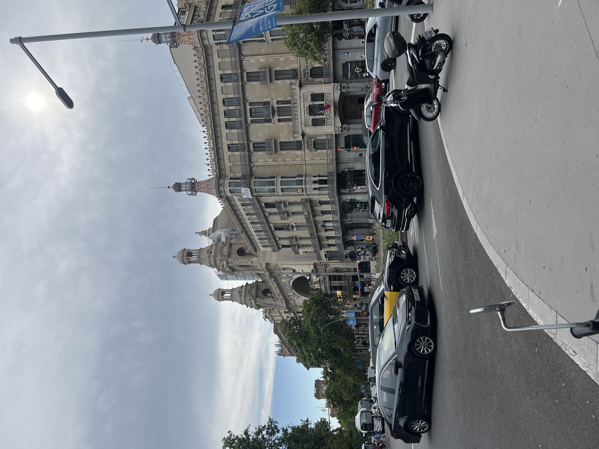 The image shows a street scene in Barcelona, Spain, with a focus on a grand building facade and busy street traffic. The building, identified by the text "COLISEUM" visible on its arched entrance, is adorned with ornate architectural details including domes, columns, and balconies. In the foreground, several cars are stopped or moving slowly at an intersection, including a black sedan, a yellow taxi, and a black SUV. A black scooter is parked on the sidewalk to the right. Pedestrians are visible on the sidewalks and crossing the street. The sky is overcast with clouds.