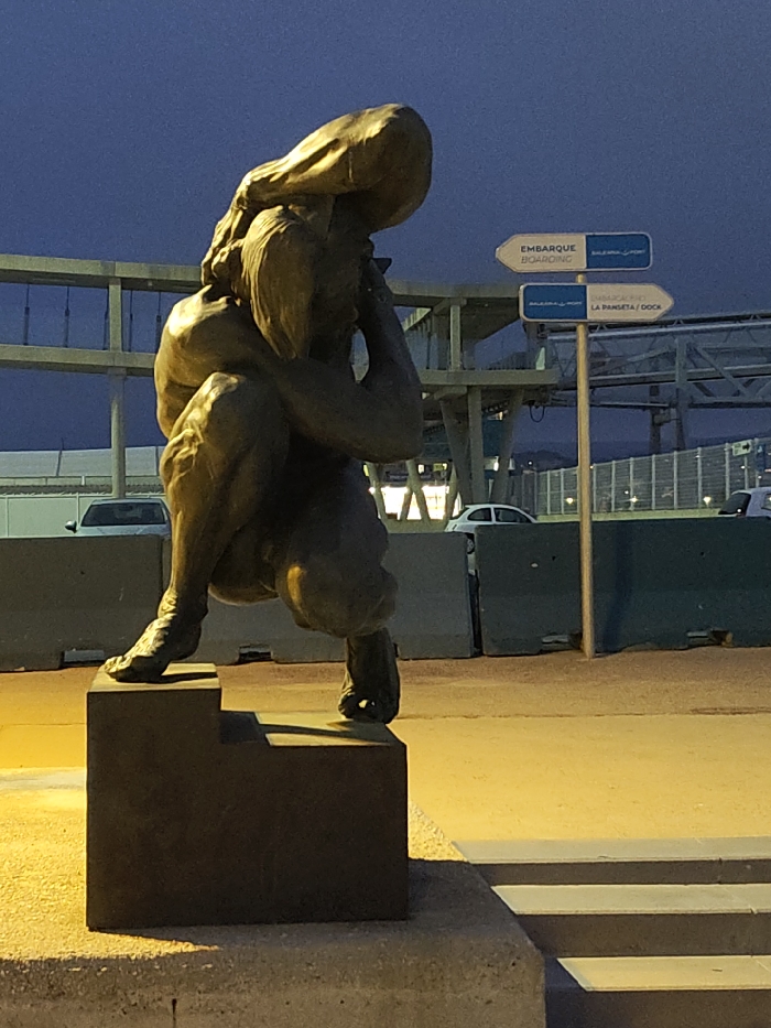 A low-angle, eye-level photograph taken during twilight or night in the port area of Dénia, Spain. The central subject is a dark bronze-colored statue of a muscular human figure in a deep squatting or crouching posture, appearing burdened. A large, rounded, sack-like object rests on the figure's back and head, with the figure's hands positioned near its face, seemingly in support or distress. The figure's left foot is flat on a dark, multi-tiered rectangular plinth, while the toes of the right foot make contact with a lower tier of the plinth.
The immediate foreground and the base of the statue are illuminated by a strong, artificial yellow light, creating pronounced shadows. Behind the statue, a grey metal signpost features two white directional arrows pointing to the right. The upper arrow indicates "EMBARQUE / BOARDING" and "BALEARIA PORT". The lower arrow points to "BALEARIA PORT" and "LA PANSETA / DOCK", confirming the setting as the Dénia port.
In the background, a row of grey concrete barriers partially obscures several parked vehicles, including at least one white car. Beyond the barriers, a complex, multi-level grey industrial structure, likely part of a ferry terminal or port infrastructure, dominates the horizon against a deep blue, fading sky. No living individuals are visible in the scene.