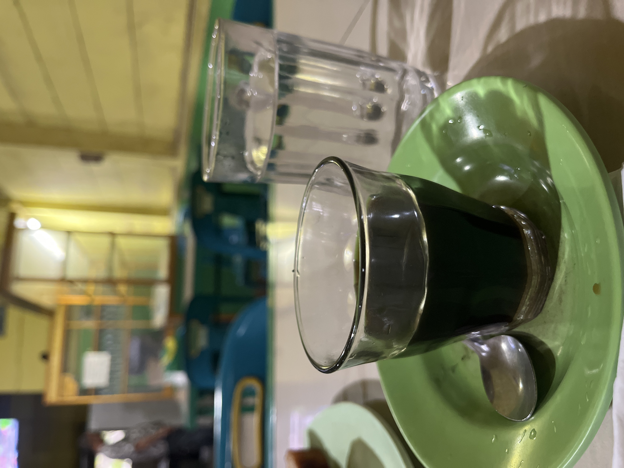 The image shows a close-up view of a table at a restaurant or cafe in Lampeuneurut Blang, Indonesia. In the foreground, a small glass of dark liquid, likely coffee, sits on a green saucer. A metal spoon rests on the saucer. To the right of the coffee glass is a tall glass filled with clear water. The table surface appears to be textured, possibly wood, with a tablecloth laid out. In the background, out of focus, are elements of the establishment's interior, including chairs, a counter with a glass display case, and a person standing, suggesting a bustling environment. The lighting is dim, with warm tones from the ambient light.