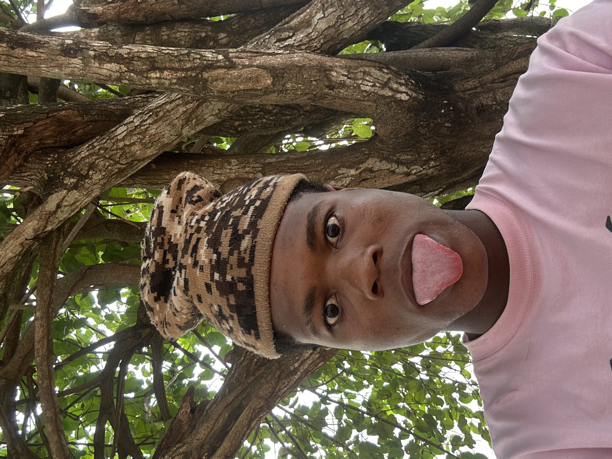 In Ihiagwa, Nigeria, a close-up, waist-up portrait captures a young Black person wearing a patterned beanie and a light pink t-shirt. Their face is turned towards the camera, eyes wide, and their tongue is stuck out. Behind them, the textured bark of a large tree is visible, with green leaves and branches forming a natural backdrop. The overall impression is one of playful expression in an outdoor setting.