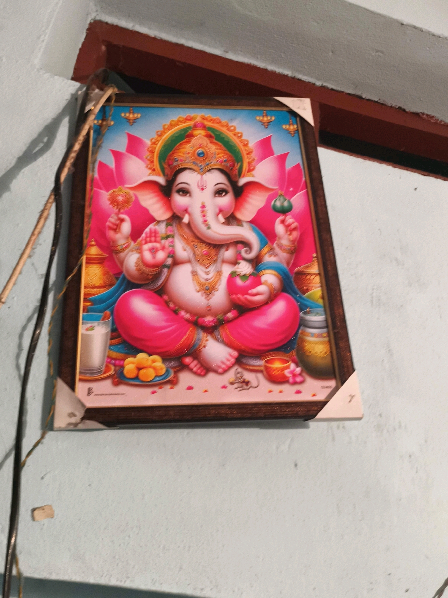 A framed devotional print of the Hindu deity Ganesha is affixed to a light blue, textured wall in what appears to be a domestic setting in Narsampet mandal, India. The print depicts Ganesha with his characteristic elephant head, seated on a pink cushion, adorned with a vibrant headdress and jewelry. He holds a lotus flower in his upper right hand and a modak (a sweet dumpling) in his lower left hand. To his left, a glass of milk and a plate of orange sweets are visible. The background features large pink lotus petals and celestial elements. The frame of the print is dark wood, and it is attached to the wall by a light-colored corner protector. A dark electrical wire runs vertically along the left side of the frame, and a thin, brown wooden dowel or stick is positioned diagonally to the left of the print. The wall itself shows signs of age and slight imperfections. The lighting suggests an indoor, possibly dimly lit, environment.