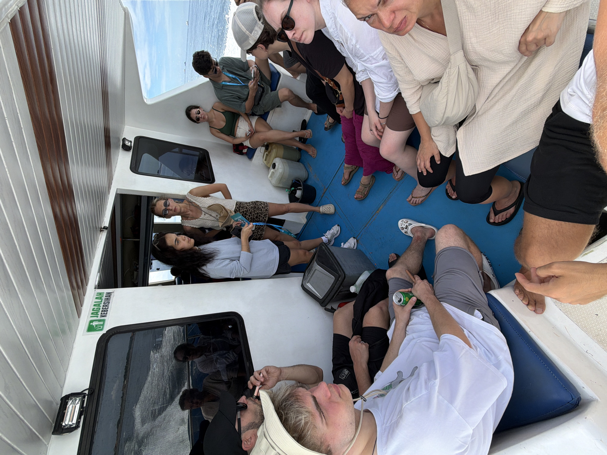 The image depicts a group of people on a boat, likely a tour boat, in or near Denpasar, Indonesia, as the green sign in the image contains the word "Jagalah Kebersihan" which means "Keep Clean" in Indonesian. There are approximately 14 people of varying ages, with most appearing to be tourists. They are sitting, lying down, or standing on the deck of the boat, which is painted white with blue accents. Some individuals are wearing sunglasses and casual clothing suitable for warm weather. One man is holding a can, possibly a beverage. The boat appears to be in motion on the water, as evidenced by the ocean visible in the background.