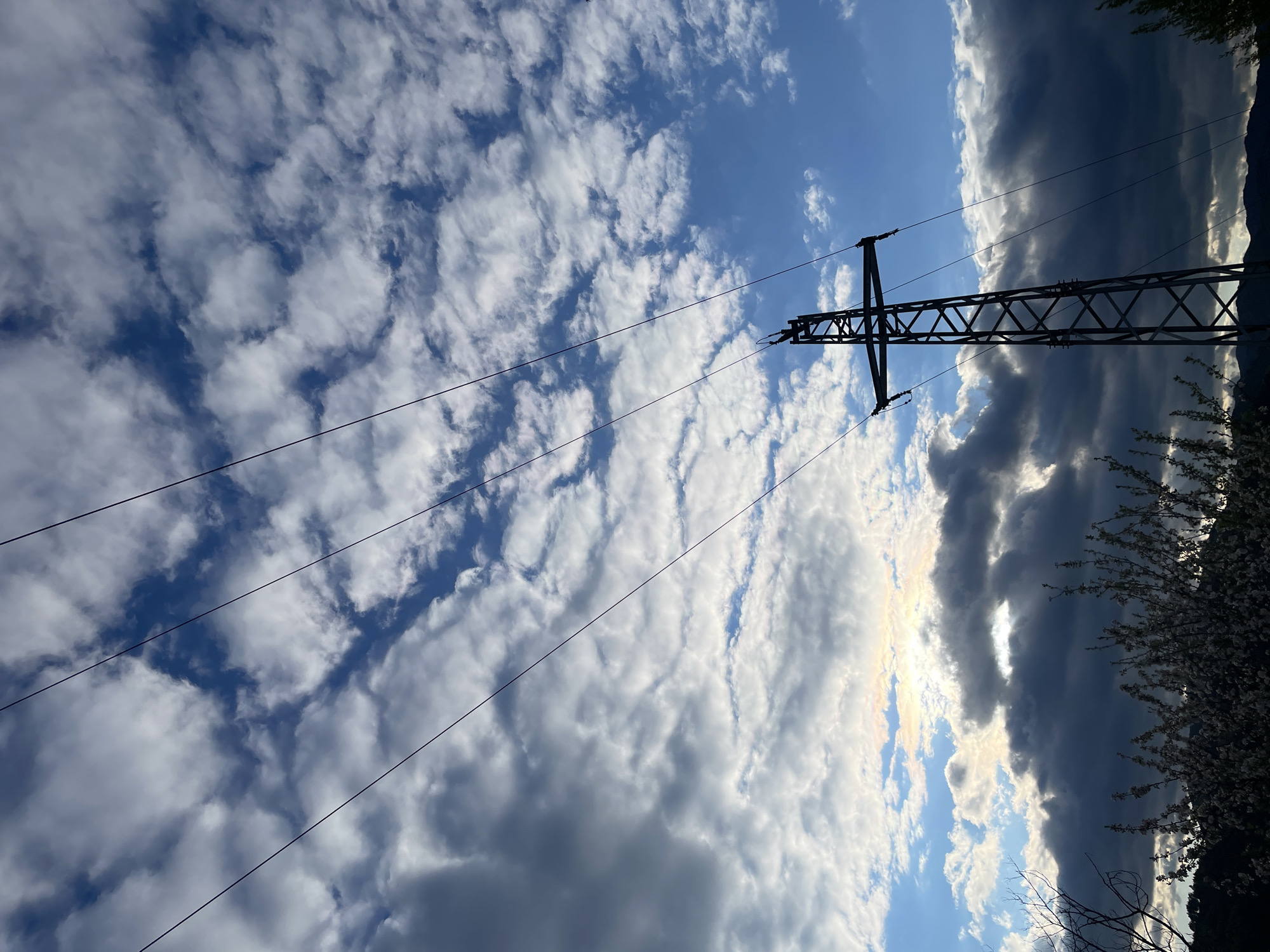 The image shows a view of the sky with white clouds and patches of blue sky, framed by an electrical transmission tower and its cables. The tower is dark in color, composed of a lattice structure, and positioned somewhat centrally in the frame. To the right of the tower, a dark, ominous-looking storm cloud is present, obscuring the sun. Vegetation, possibly trees or bushes, are visible on the right side of the frame, near the storm cloud. The photograph is taken from a low angle, looking upwards. This image was taken in Zlatograd, Bulgaria, in reference to your instructions.