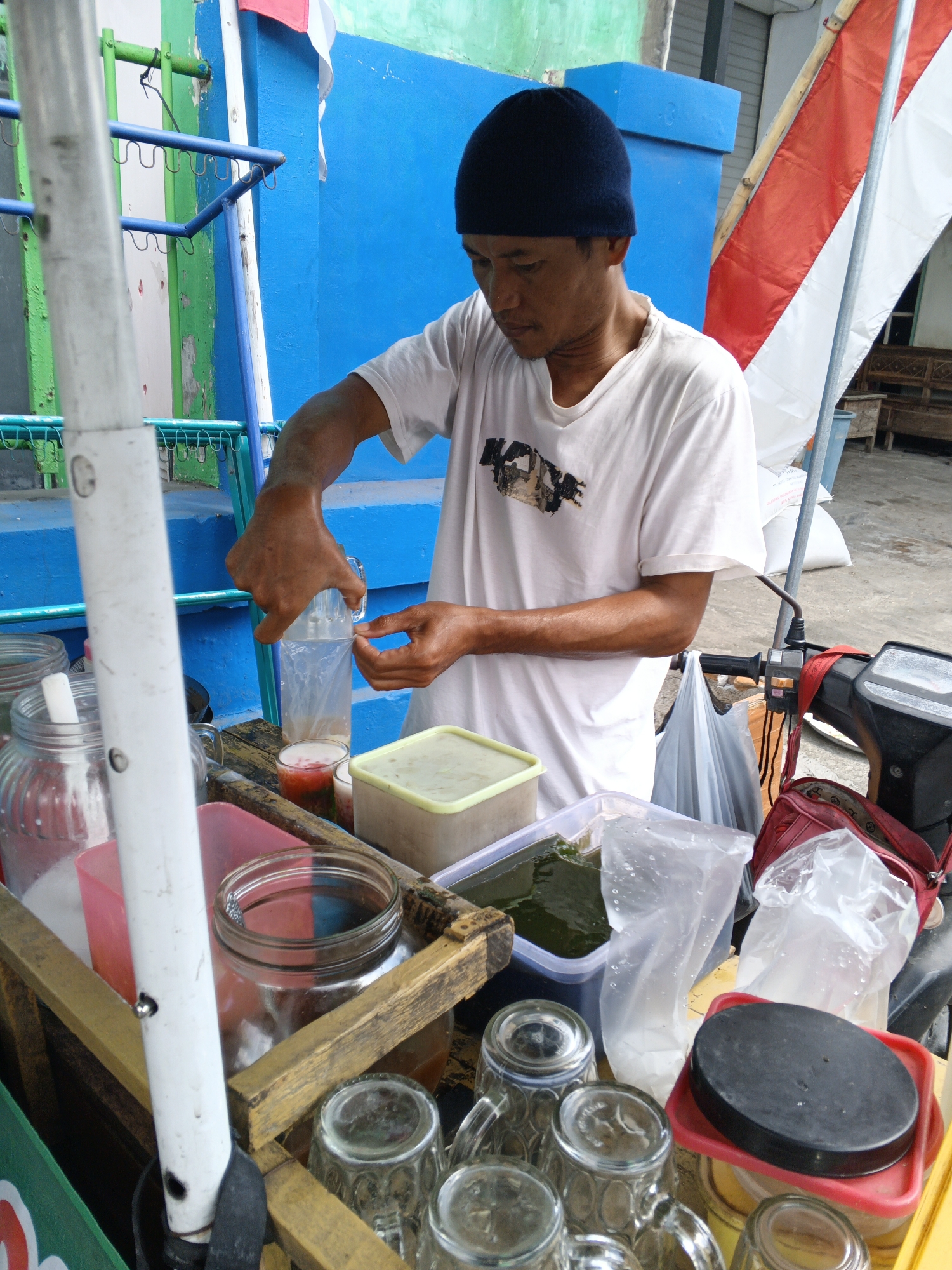 The image depicts an outdoor scene, likely a street vendor in Padang, Indonesia. A man wearing a dark beanie and a white t-shirt is in the process of preparing a drink. He is holding a clear plastic container with a handle, pouring liquid into a glass that already contains a colorful beverage. Several other containers and glasses are visible on a wooden cart.
The cart is filled with various items related to drink preparation and service:
* A large, clear glass jar with a white lid and a white straw inside.
* A red plastic container.
* A light green rectangular plastic container with a light green lid, containing a green substance, possibly a jelly or herb.
* Several clear glasses, some with handles, arranged on the cart.
* A large, clear plastic bag filled with white material.
* A red bag is also visible on the cart.
In the background, a brightly painted blue wall stands out. A red and white flag, likely the Indonesian flag, is draped from a pole. A portion of a motorcycle is visible on the right side of the image, with its handlebars and a side mirror. The ground appears to be paved. The overall setting suggests a typical street-side food or beverage stall.