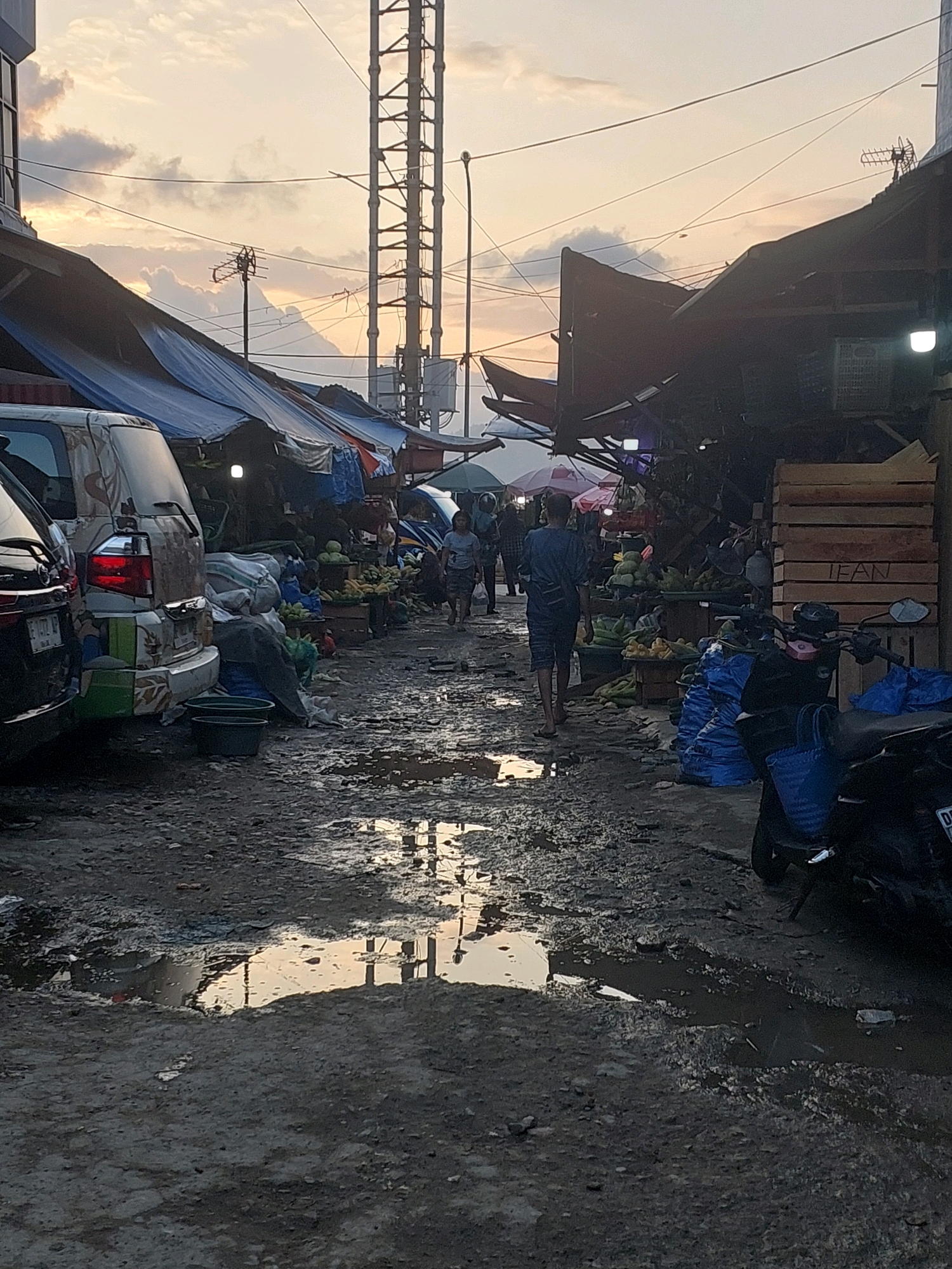 The image depicts a street market scene in Ambon, Indonesia, likely during the late afternoon or early evening, given the warm, diffused light and overcast sky with some clouds tinged with orange. The ground is muddy and uneven, with several puddles reflecting the sky and surrounding structures.
To the left, a white van with colorful decals is parked, its rear visible. In front of it, several large sacks are piled up, some covered with a gray tarp. Two dark green basins sit on the ground next to the sacks. Further down the lane, more sacks, some blue, are visible, along with various produce, including corn and what appear to be coconuts or similar fruits, displayed in crates and on the ground. A motorbike is parked on the right side of the lane.
On the right side of the lane, several market stalls are set up with awnings made of blue tarpaulin. One stall has wooden crates stacked, with the word "IEAN" scrawled on them. Brightly lit bulbs hang from some of the stalls, illuminating the goods.
Several people are present in the market. A person in a blue shirt and shorts walks away from the camera down the lane. Further in the distance, two people are walking towards the camera, and other figures are visible amongst the stalls. A tall, lattice-like tower, possibly a telecommunications mast, stands prominently in the background, a common sight in urban Ambon. Electrical wires crisscross the sky above the market. The overall atmosphere suggests a working market, with goods being sold and people going about their business amidst the somewhat dilapidated and wet conditions.