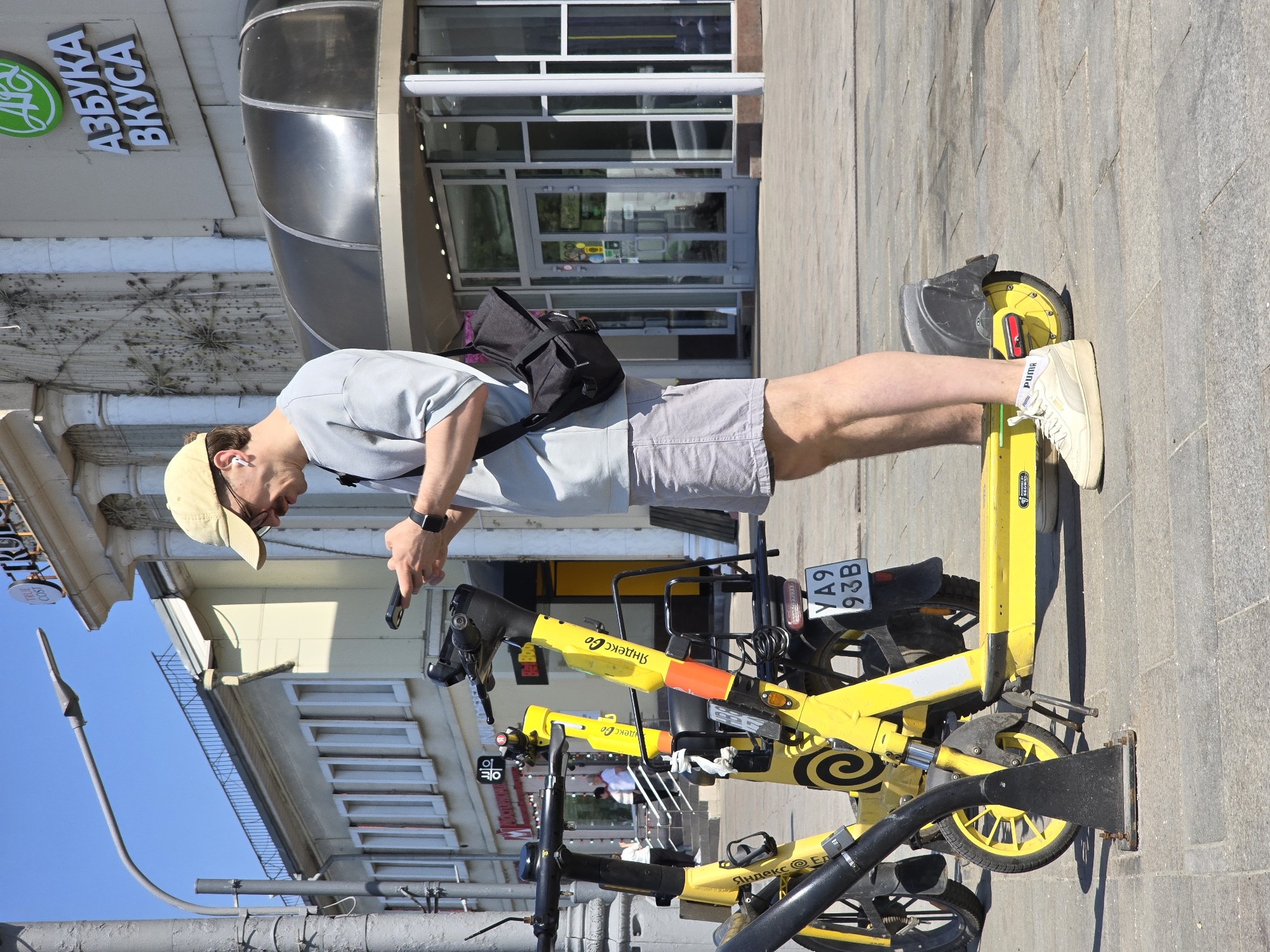 A medium, eye-level, medium-shot photograph shows a man in Moscow, Russia, standing next to three parked, yellow, electric scooters.
The man is facing the scooters and is angled toward the camera left. He wears a tan baseball cap, sunglasses, a gray T-shirt, gray shorts, white sneakers with a Puma label, and a black bag slung over his left shoulder. He appears to be unlocking a scooter with a smartphone in his right hand and is wearing a black smartwatch on his left wrist.
Three electric scooters are parked to the man's right. The scooters have black handlebars and black fenders. The scooters have “Yandex Go” written in Cyrillic on the frame. The scooter closest to the camera has a license plate reading “YA9 93B.” All scooters have black metal bars affixed to their bases to prevent them from being moved.
Behind the man and the scooters is a building with a light gray facade. There is a sign reading “Азбука Вкуса” and a green circular logo. There is a bus shelter attached to the building entrance.
In the background is a blue sky. A tall, gray light post is visible on the left.