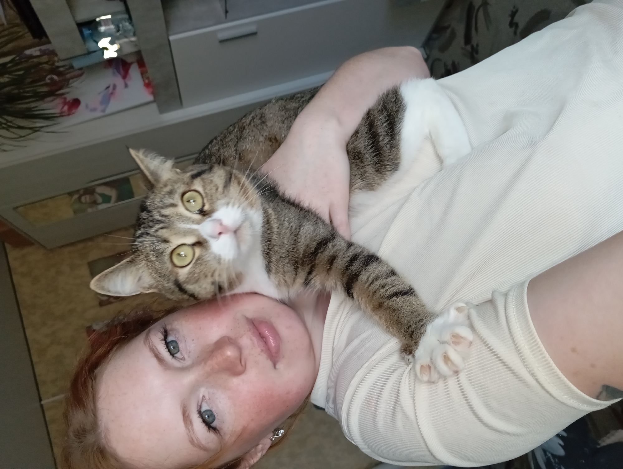 A person is holding a cat. The person has fair skin, freckles, and blue eyes. The cat is brown and white tabby. They are in an indoor environment, possibly a home. The location is unknown.