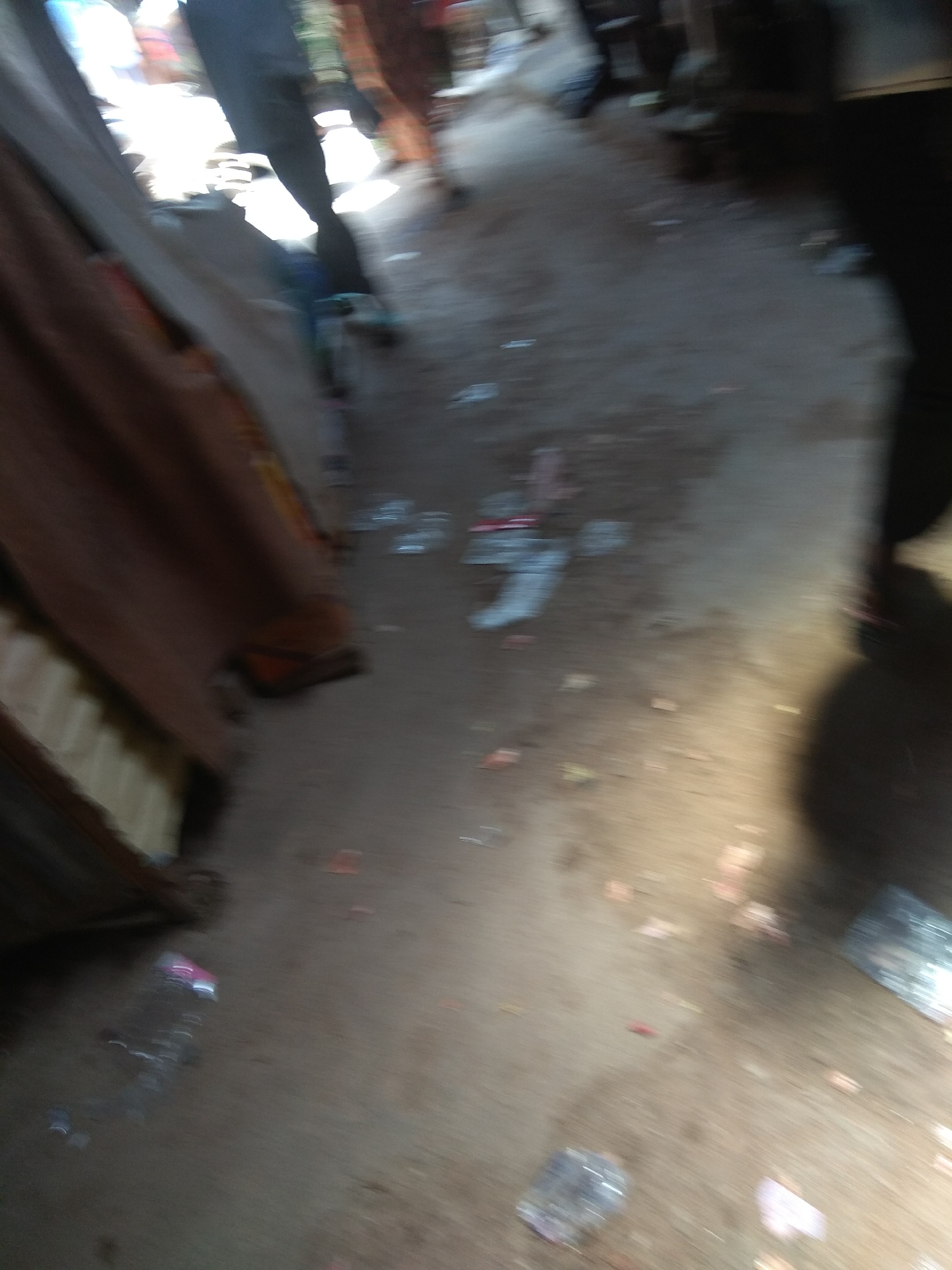 The image depicts a dusty, unpaved outdoor area that appears to be a market or thoroughfare in Jalingo, Nigeria. The scene is characterized by a sense of disarray, with visible litter on the ground, including plastic bags and wrappers. The lighting suggests it is daytime, with harsh sunlight casting strong shadows. The ground is a mixture of dirt and what looks like fine gravel.
In the foreground, a partially visible plastic water bottle lies on the ground. To the left, a tall, brown, fabric-covered structure, possibly a stall or awning, dominates the left side of the frame, obscuring further details of the immediate surroundings.
Further into the scene, there are blurred figures of people, suggesting movement and activity. One figure in the background appears to be wearing dark trousers. The overall impression is one of a busy, somewhat neglected urban environment. The motion blur in the image indicates that either the camera or the subjects were in motion during the capture.