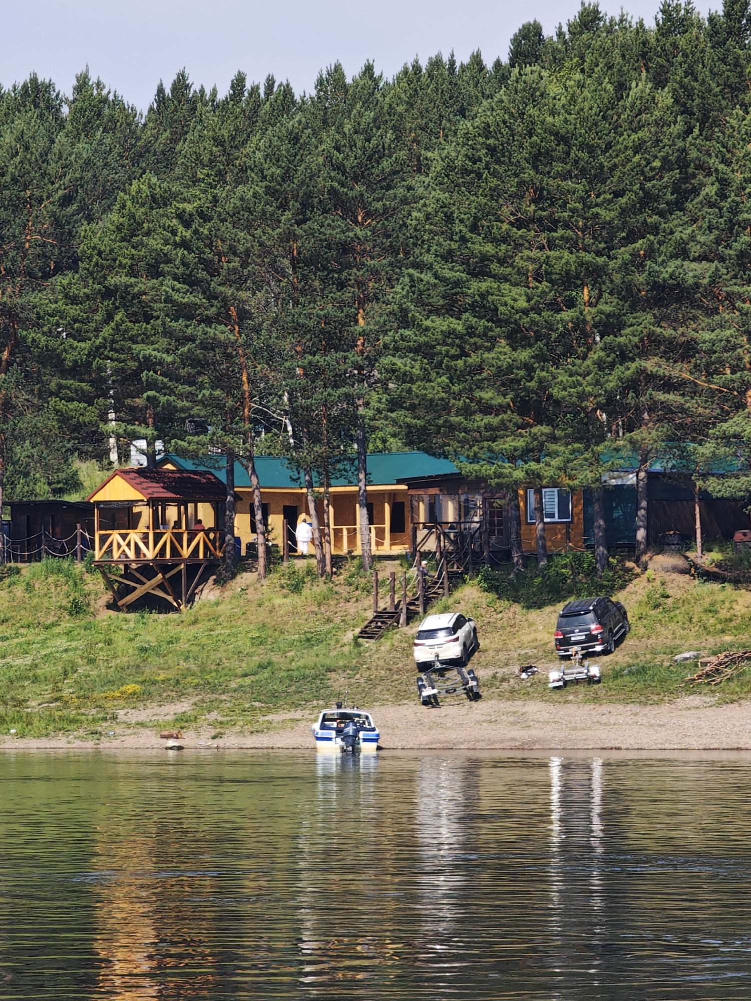 The image depicts a waterfront scene in Podporozh'e, Russia, featuring a wooded hillside with a few buildings and vehicles near the water's edge. A dense forest of tall pine trees forms the backdrop, extending across the top of the frame. On the slope, a cluster of wooden structures with yellow walls and green roofs are visible. One building has an elevated wooden porch with a red roof.
In front of the buildings, on a sandy and grassy incline, are two vehicles: a white SUV with a trailer and a black SUV with a trailer. A small white and blue motorboat is docked at the edge of the water in the foreground. The water itself is calm, reflecting the sky and the surrounding greenery. The overall atmosphere is that of a rural retreat or a riverside settlement.