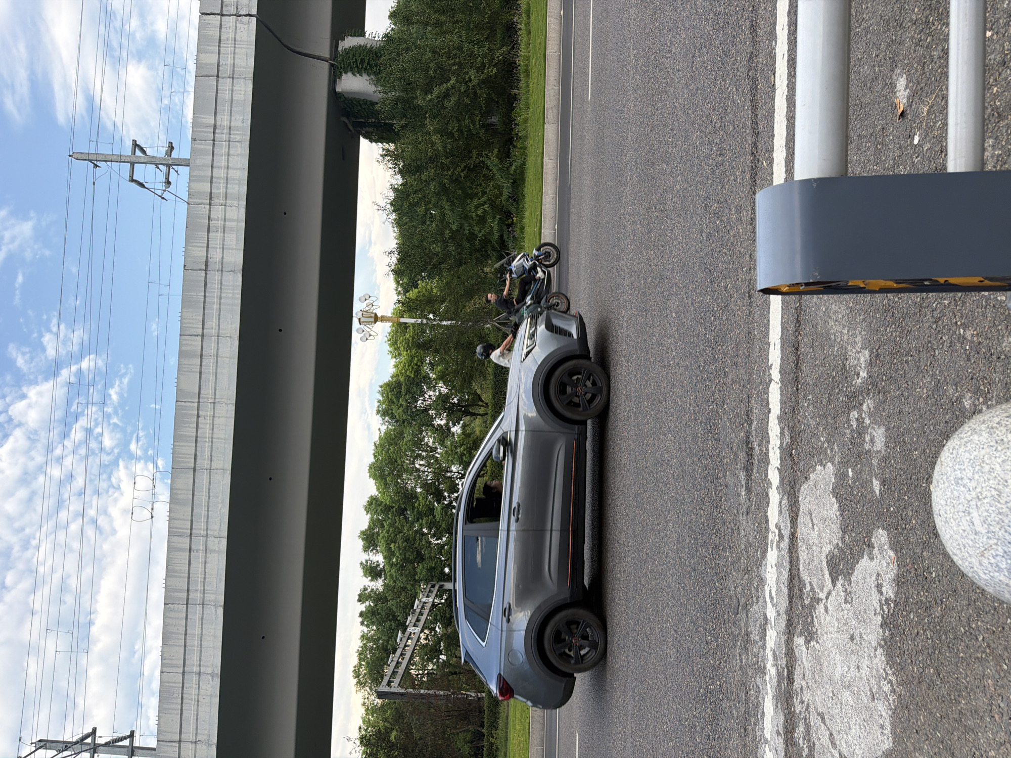 A grey SUV is driving on a road in the city of Jiangjin, China, under a large overpass. A person is visible in the driver's seat. Two people on motorcycles are also driving on the road. The environment consists of a road, trees, and a cloudy sky. A traffic camera pole is visible on the left side of the road.