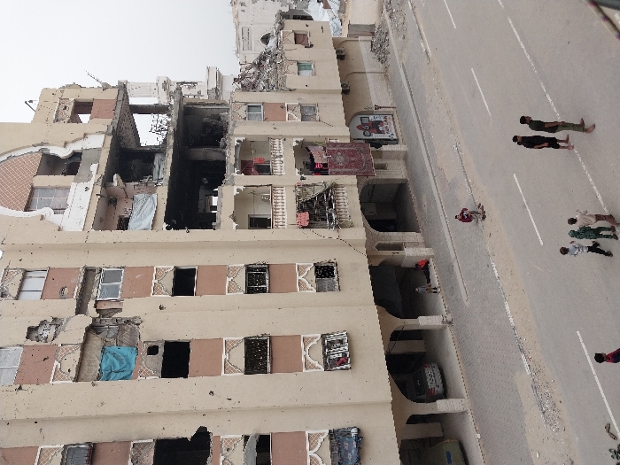 A tan building reveals extensive damage from bombing or shelling. Several floors are gutted with rooms exposed, debris visible, and structural damage apparent. On the street, several people walk along the road in small groups. A car is parked in an alcove on the ground floor of the building. The scene conveys destruction and the presence of civilians in a war-torn environment.
