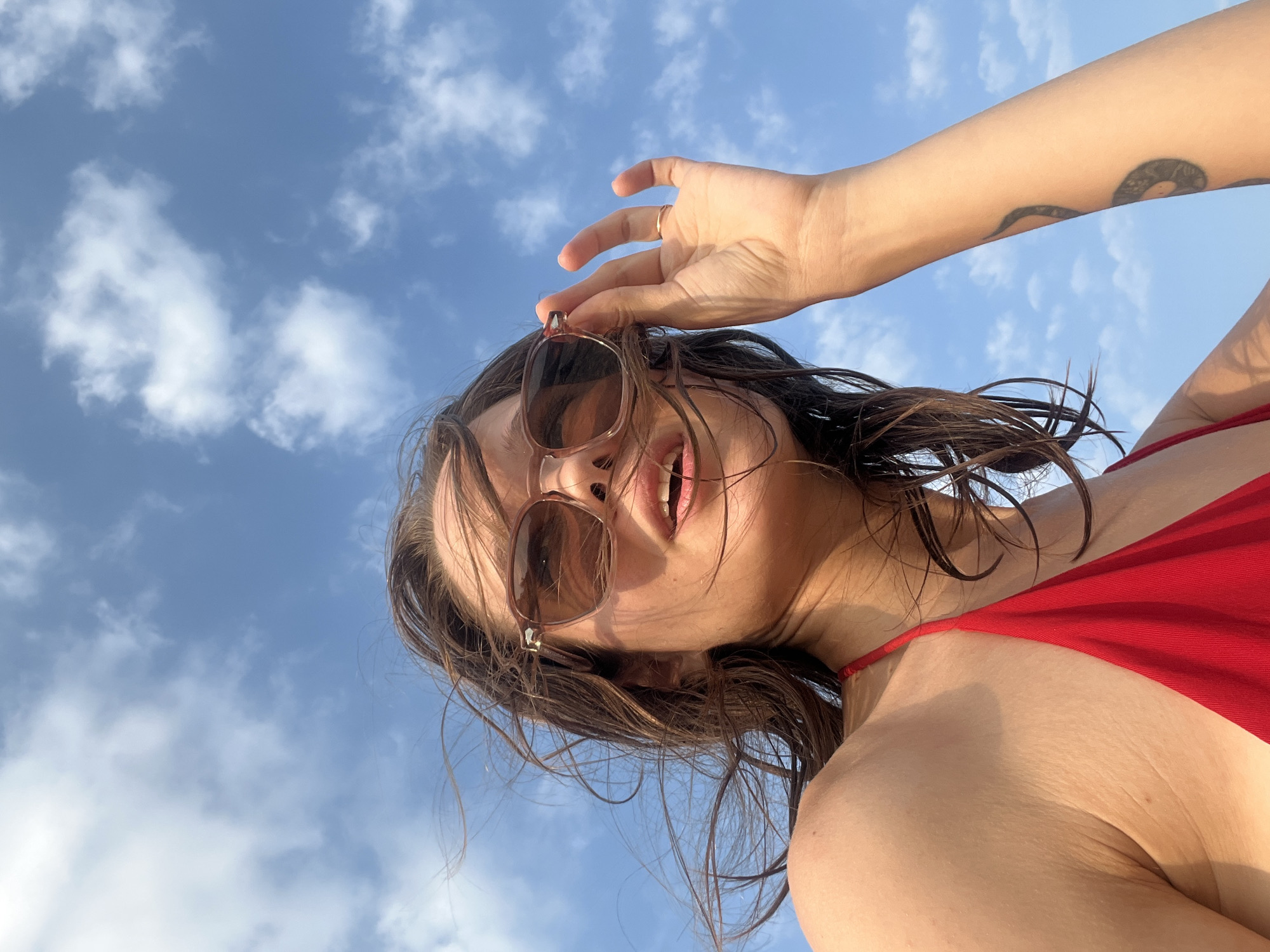 A low-angle, close-up shot features a person, likely female, looking down towards the camera against a bright blue sky with scattered white cumulus clouds. The subject's face is partially framed by loose, dark brown hair, which appears slightly damp or windswept. She wears brown-tinted sunglasses that obscure her eyes, and her mouth is open, suggesting a smile or laugh.
She is dressed in a red garment, likely a swimsuit or bikini top, visible across her chest and shoulder. Her right arm is raised, with her hand positioned near her temple or hair, and a simple gold-colored ring is visible on one of her fingers. On her left arm, a dark, stylized tattoo with a serpentine or abstract design is clearly visible against the sky.
The lighting is natural and bright, indicative of a sunny daytime. The low perspective creates an uplifting and open feel, emphasizing the vast sky above. The scene conveys a relaxed, casual outdoor moment. The context provided indicates the location is Nam Nha Trang, Vietnam. There is no visible text in the image.