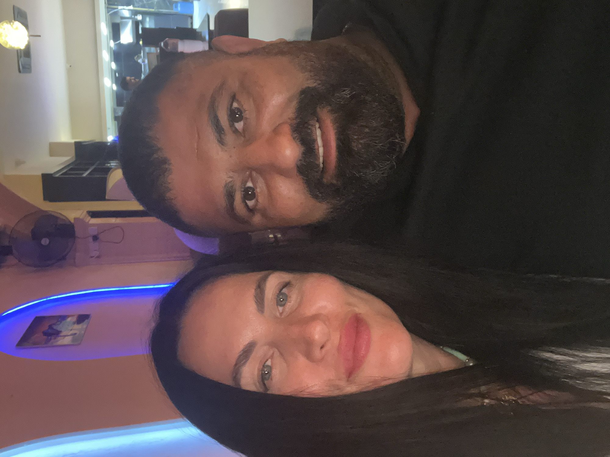 The image is a close-up portrait of two smiling individuals, a man and a woman, presented rotated 90 degrees clockwise. Their heads and shoulders fill most of the frame, suggesting a selfie-style photograph.
On the left side of the frame (when viewed upright), a man with a dark complexion, a full dark beard, and short dark hair is looking directly at the camera with a broad smile, revealing his teeth. He is wearing a dark-colored top.
On the right side of the frame, a woman with a lighter complexion, long dark hair, and light makeup including pink lipstick, also looks directly at the camera with a subtle, relaxed smile. She appears to be wearing a light-colored top, and a piece of jewelry, possibly a ring or bracelet, is faintly visible on her wrist.
The setting is an indoor establishment, likely a restaurant, bar, or lounge, indicated by the artificial lighting and decor. In the background, vibrant neon blue strip lights illuminate a curved ceiling or wall section behind the woman. A distinct yellow, spherical light fixture is visible behind the man. Other background elements include faint dark shelving units or cabinets, a small framed picture on the wall, and a black fan. The predominant artificial lighting suggests the photo was taken at night or in a dimly lit interior. Both individuals convey a sense of contentment and relaxation. This scene is located in Kyrenia, Northern Cyprus / Cyprus. No visible text is present in the image.