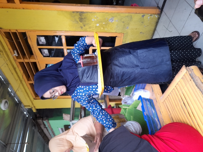 A woman in a blue hijab and polka-dot top stands behind a yellow tray holding a jar. She wears glasses and is positioned near a yellow building. Below, other women are present, one in a light tan hijab and another in red. Blue plastic crates and bamboo fencing surround them. The setting appears to be a market or street-side vendor location.