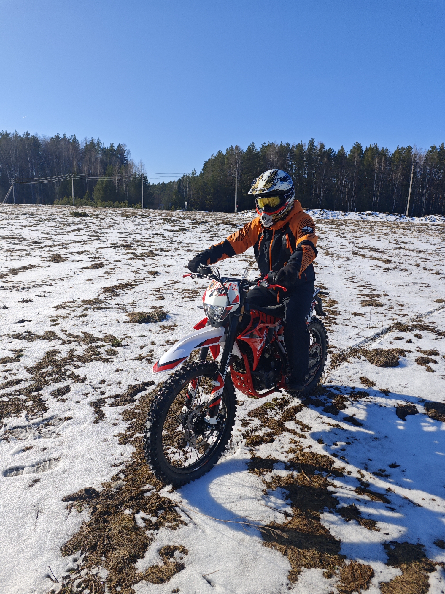 The image depicts a person riding a red and white Beta dirt bike in a snow-covered field. The rider is wearing a full-face helmet with goggles, an orange and black jacket, and black pants. The field is a mix of snow and patches of brown grass. In the background, there is a line of trees under a clear blue sky. There is a power line crossing the background. The location is near the city of Прикамский, Russia.