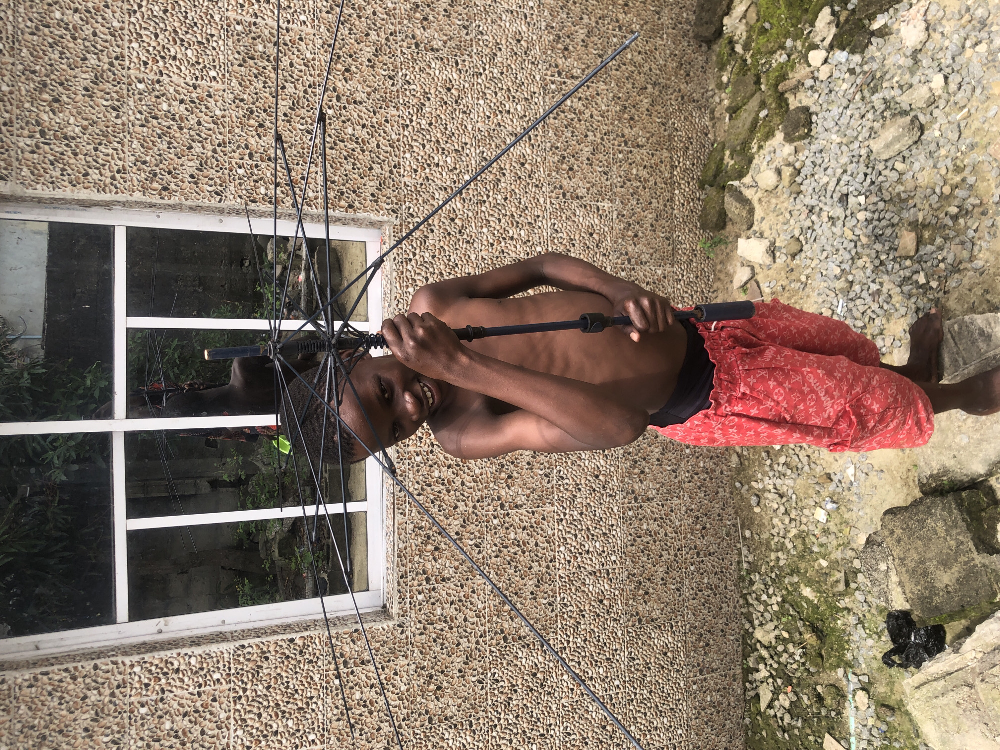 A young boy, appearing to be around ten years old, stands shirtless and smiles at the camera, holding an open black umbrella by its handle. He is wearing red patterned shorts with a black waistband. The background consists of a textured wall made of small, light brown and beige pebbles, with a white-framed window above. Through the window, a dense green foliage and some gray structures are visible, suggesting an outdoor or semi-outdoor setting. The ground in front of the wall is covered with small gray gravel and some larger stones, with patches of moss and green growth. A black plastic bag lies on a concrete block to the left of the frame. The overall impression is a candid shot of a child in an urban environment, possibly in Ado, Nigeria, as indicated by the context.
