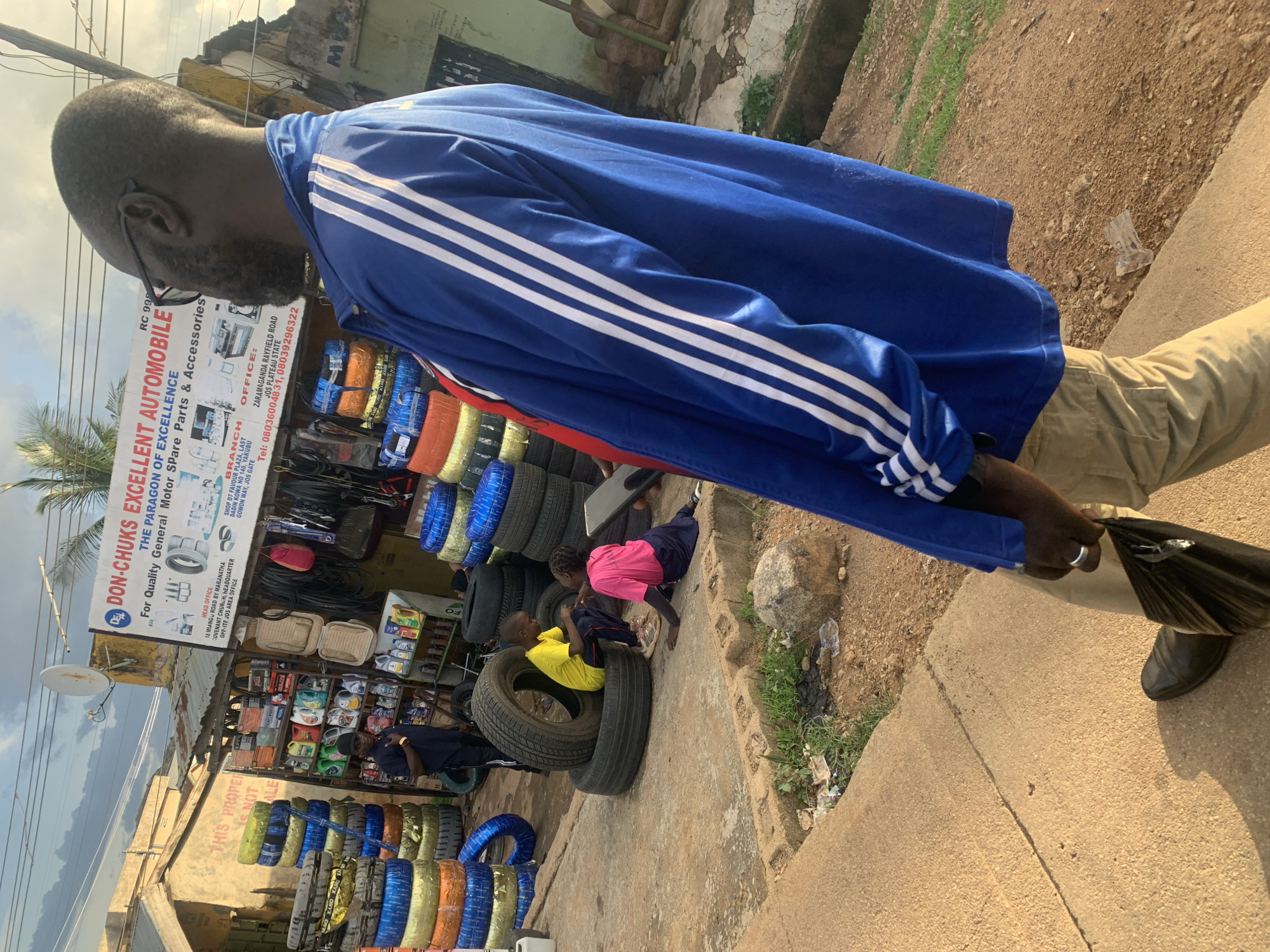 This image shows a scene on a street in Jos, Nigeria, likely during the day given the lighting. The foreground features a man wearing a blue tracksuit with white stripes and beige pants, walking and carrying a black plastic bag. He is wearing glasses and has a beard. Behind him, a store is visible, displaying various automotive spare parts and accessories, including numerous tires of different colors stacked and arranged. A sign for "DON-CHUK'S EXCELLENT AUTOMOBILE" is prominently displayed, listing their office and branch locations in Jos. In the mid-ground, two children are sitting on tires. One child is wearing a yellow shirt and the other a pink shirt, appearing to interact or play. The overall setting is an outdoor market or roadside shop, with a gritty, urban feel, including some debris on the ground. The weather appears to be overcast.